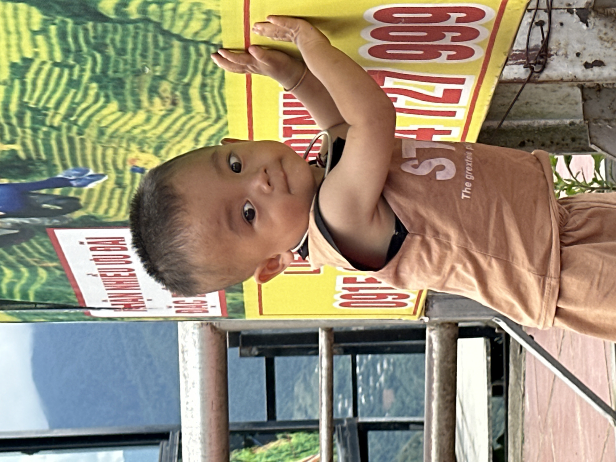 In the city of Tu Le, Vietnam, a young East Asian child with short, dark hair is positioned in the foreground, facing slightly right of the viewer with an inward gaze. The child wears a light brown sleeveless top displaying "STA" and "The greate" (partial) in white text, over a dark undergarment, and matching shorts. A thin, light-colored bracelet is on the child's right wrist, and a dark strap is around the neck. Both of the child's hands are pressed against a large, vertical sign that is predominantly yellow with a green upper section. The green section depicts a pattern resembling terraced fields and a blue human figure's legs. The sign features Vietnamese text in white on a red horizontal bar: "NHAN NHIEU Uu DAI," and red numbers on the yellow background, including "0915" (partial) on the left and "727 999" on the right. The sign is mounted on weathered metal posts. In the mid-ground to the left, a rusted metal railing runs horizontally. Behind the railing and the sign, hazy blue mountains and distant green foliage are visible, indicating an elevated outdoor setting. The ground directly beneath the child is a reddish-brown paved surface.