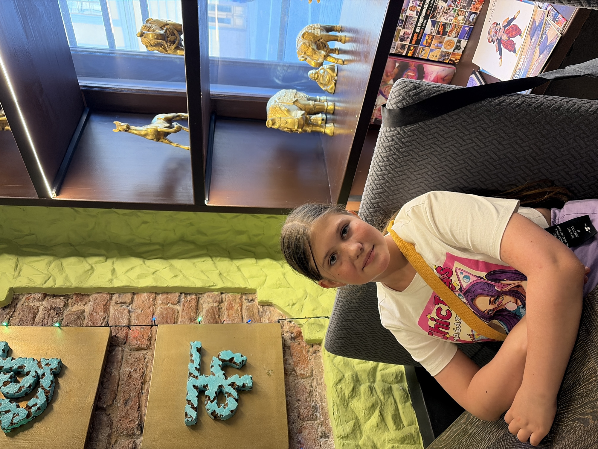 In a cafe in Saint Petersburg, Russia, a young girl with blonde hair pulled back is seated at a table. She is wearing a white t-shirt with a graphic of a character with purple hair and headphones, and a yellow strap across her chest. Her arms are crossed, and she is looking at the camera. Behind her, a wall is decorated with textured lime-green paint and brickwork, featuring two gold plaques with turquoise ornamental designs. To the right, a dark wood shelf unit displays various golden figurines, including a horse, elephants, and a seated figure, likely Buddha. The shelves are illuminated by LED lights. Books and magazines are stacked on a lower shelf. The overall atmosphere suggests a cultural or themed establishment.