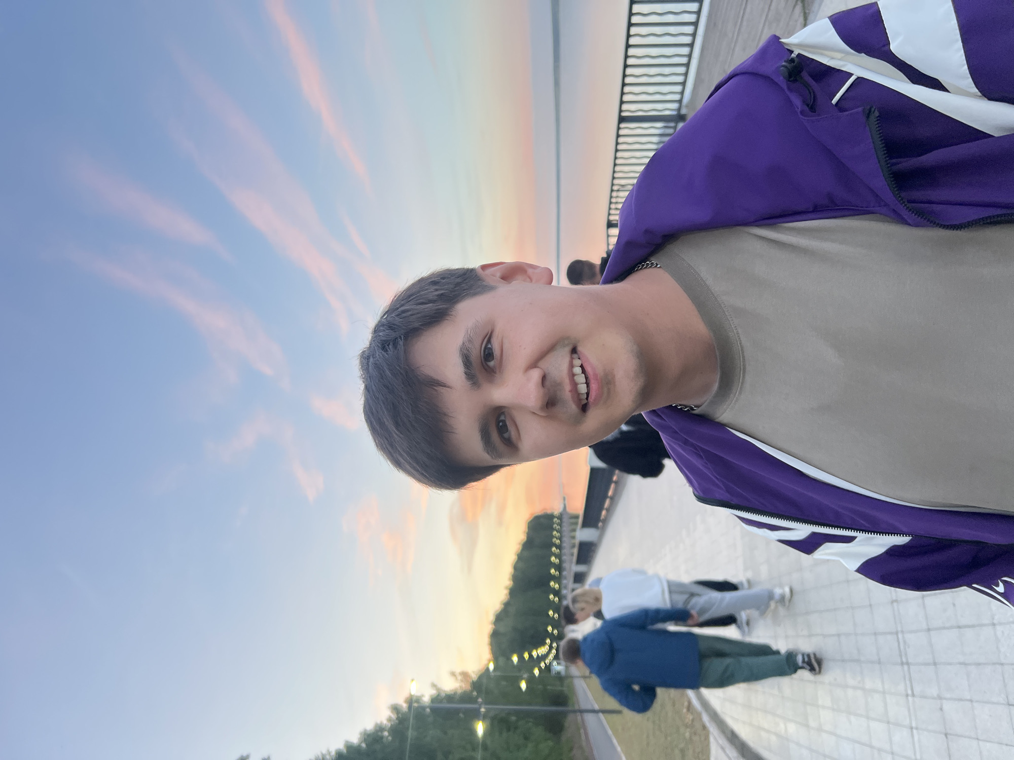 A young man takes a selfie on a paved waterfront promenade in Chaikovsky, Russia, during what appears to be sunset. The image is oriented vertically but displayed rotated 90 degrees counter-clockwise. The man is smiling broadly at the camera, revealing his teeth, and has short dark hair. He is wearing a light beige t-shirt and a vibrant purple and white windbreaker jacket, with a silver chain visible around his neck.
The background features a serene sky illuminated by the soft, warm light of either sunrise or sunset, displaying pastel shades of blue, pink, and orange, with some streaky clouds. Below the sky, a large body of water, likely the Kama River or Votkinsk Reservoir, stretches towards the horizon. A black metal railing borders the promenade, separating it from the water. On the distant shore or hillside, green trees and several streetlights or decorative lights are visible, indicating an established public area.
The man is standing on a light-colored, patterned paved surface. Further down the promenade, two other people are partially visible, appearing to walk or stand casually. One is wearing a light-colored hoodie and pants, and the other a dark blue jacket and green pants. The scene suggests a pleasant, leisurely evening activity in a scenic outdoor location. The man's expression conveys happiness and contentment. No discernible text is visible within the image.