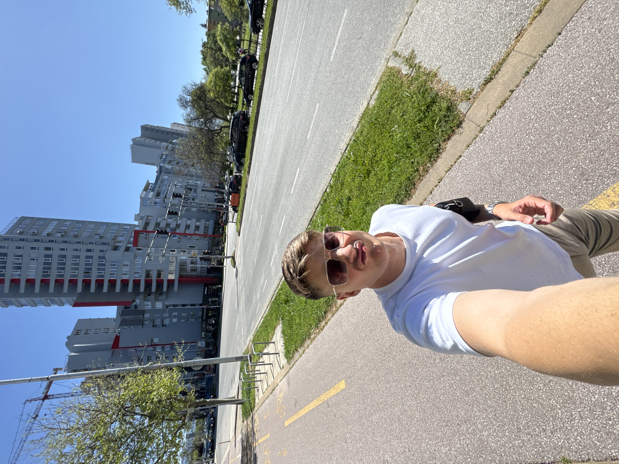A medium-shot selfie depicts a fair-skinned man wearing sunglasses and a white t-shirt, possibly sticking his tongue out slightly. He stands on a paved path with yellow directional lines. To his right is a strip of grass and a road with parked cars. Beyond the road, in the background, stand several tall, modern buildings constructed with red and grey tones. A construction crane is visible near the buildings. The sky is clear and blue. The location appears to be in the city of Plovdiv, Bulgaria, based on the architectural style of the buildings.