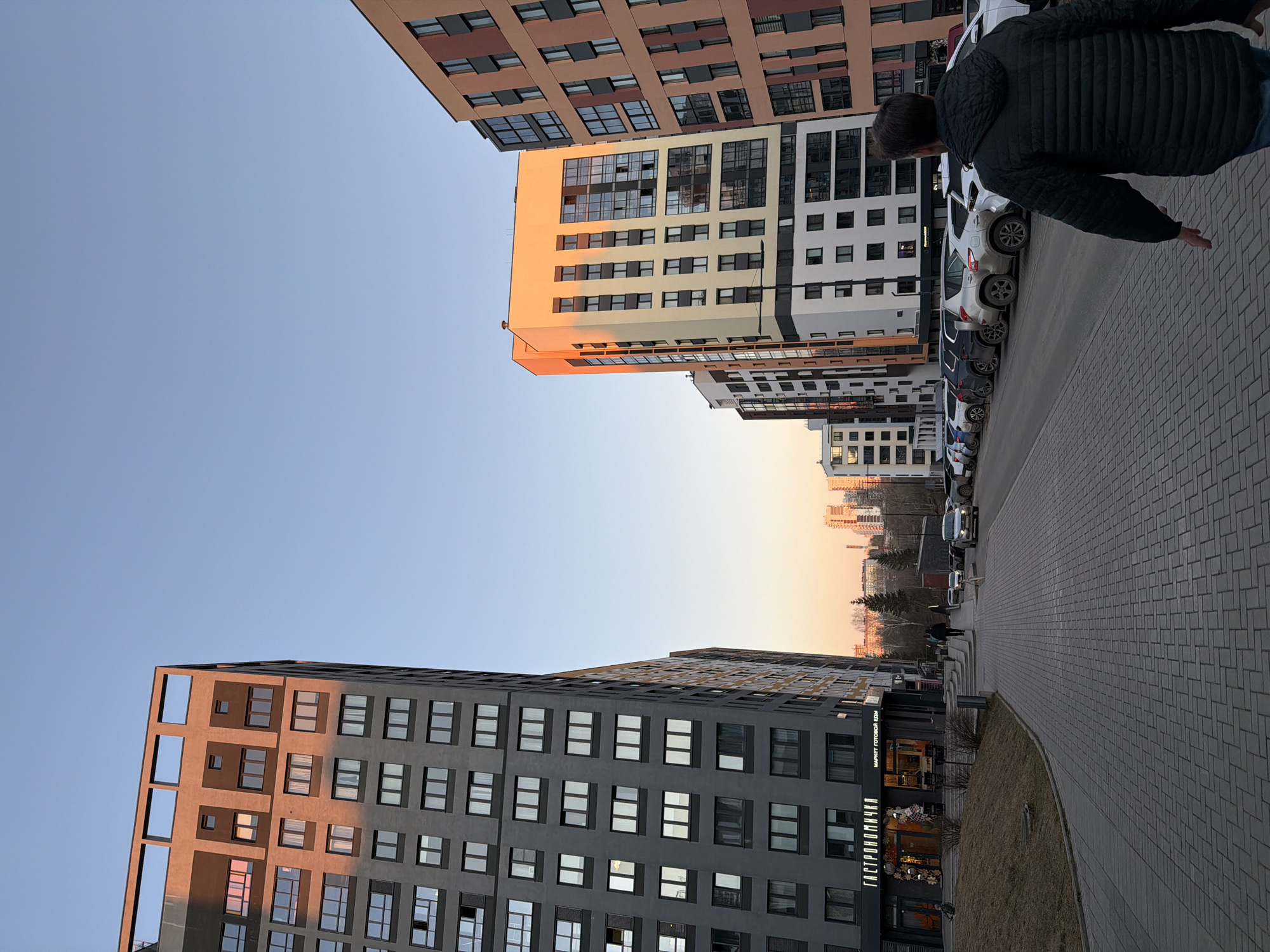 An eye-level outdoor shot shows a street scene in Izhevsk, Russia. Two high-rise buildings frame the sides of the image, leading the viewer's eye down the street. The building on the left is a dark gray, modern structure with evenly spaced windows. The building on the right is multi-colored with a mix of beige, brown, and white sections.
A line of parked white cars runs parallel to the building on the right. A man wearing a dark quilted jacket is partially visible in the top right corner, walking along the paved sidewalk. The sidewalk is made of gray interlocking bricks.
In the distance, more buildings and trees are visible under a warm, golden sky, suggesting the time is either sunrise or sunset. The sky above the buildings is a clear, pale blue. A store sign, "ГАСТРОНОМИЧКА," is visible on the lower portion of the building on the left.