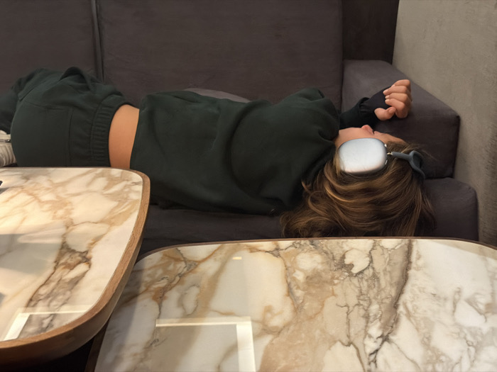 An individual is depicted lying on their back on a dark grey fabric sofa in an indoor setting in Mexico City, Mexico. The person, whose gender is not definitively ascertainable but appears to be female, is wearing dark green athletic wear consisting of a long-sleeved top and sweatpants, exposing a portion of their midriff. Light-colored over-ear headphones are worn over their brown hair. Their left arm is bent, with the hand resting near their head on the sofa cushion. In the foreground, two coffee tables with white and brown marble tops are partially visible, one featuring a light wooden edge. The background consists of a plain wall. The individual appears to be resting or relaxing.