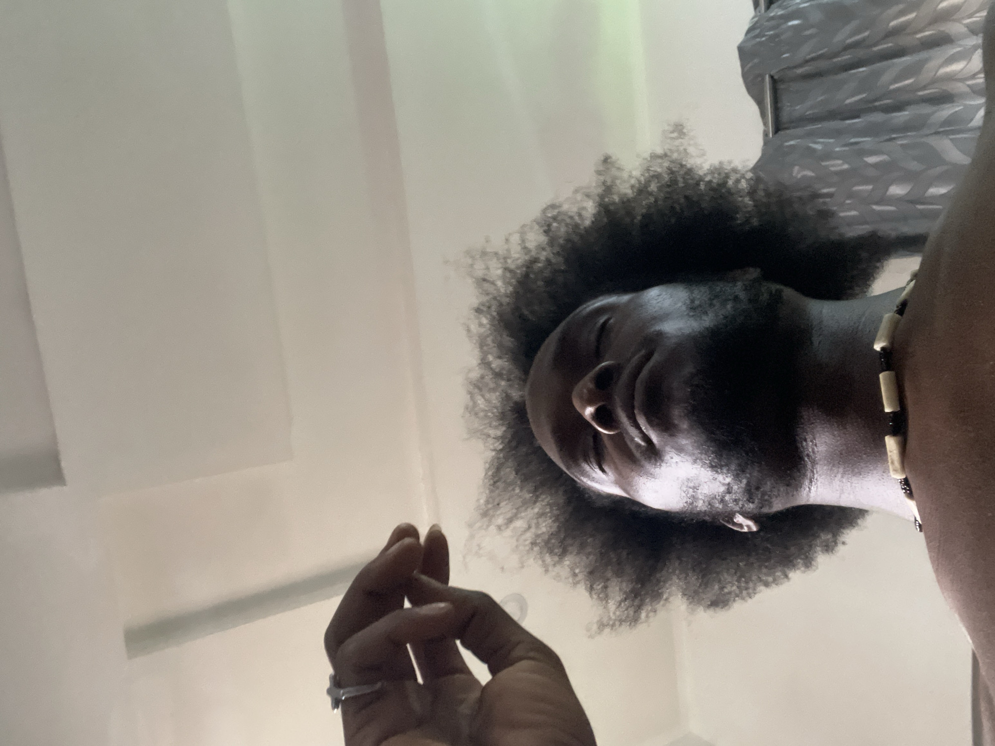 A low-angle shot of a Black male with dark curly hair and a beard, viewed from below. He has a nose piercing in his left nostril and a beaded necklace. A hand with a ring on the ring finger is in the lower left corner of the image. The background consists of a white ceiling and gray curtains in the upper right corner. The photograph appears to have been taken indoors. The location is unknown and there is no evidence that the image was taken in the city of Kuchingoro, Nigeria.