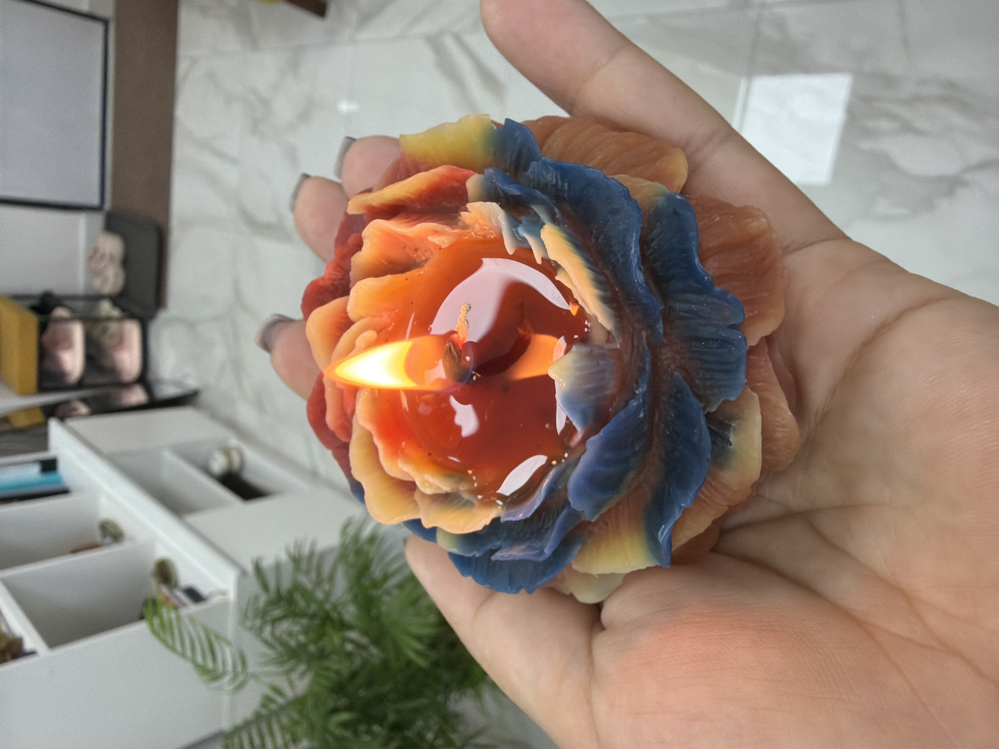 A hand holds a multi-colored rose-shaped candle that is lit and melting. The candle has petals that transition from blue at the base to orange and red at the top, where the flame is located. The flame is bright yellow and orange, with a small amount of melted wax pooling around the wick. The background is blurred but appears to be an indoor setting with a tiled floor and some furniture, possibly a shelf unit with plants and decorative items. The image is a close-up, focusing on the candle and the hand. The specific location of Petropavlovsk-Kamchatsky, Russia, cannot be determined from the visual information in the image.