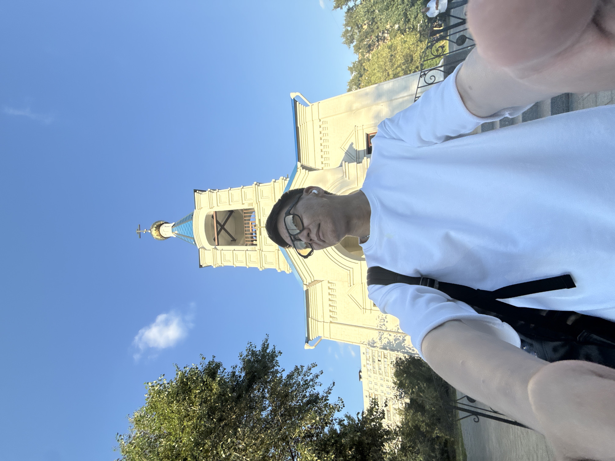 The image is a low-angle, selfie-style shot taken outdoors on a sunny day, with a clear blue sky. In the foreground, a person wearing a white t-shirt, sunglasses, and white wireless earbuds is visible, holding the camera out to capture the scene. A black backpack is slung over their left shoulder. The person is looking upwards and to the right.
Behind the person, a light-colored building with a blue roof and a golden dome topped with a cross is prominently featured. This appears to be a church or a similar religious building. The architecture is classical, with arched windows and decorative elements. To the left of the building, a large green tree with dense foliage is visible. In the background, a tall apartment building with numerous windows can be seen. A low black metal fence with ornate scrollwork runs along the lower part of the frame, separating the viewer from a paved walkway or path.
The specific location is identified as the Church of the Nativity of the Blessed Virgin Mary in Dolgoprudny, Russia. The building's architecture, including the dome and cross, aligns with common designs of Russian Orthodox churches. The presence of a modern apartment building in the background suggests an urban or suburban setting, typical for many churches within Russian cities. The overall atmosphere is bright and clear, indicative of a pleasant daytime.