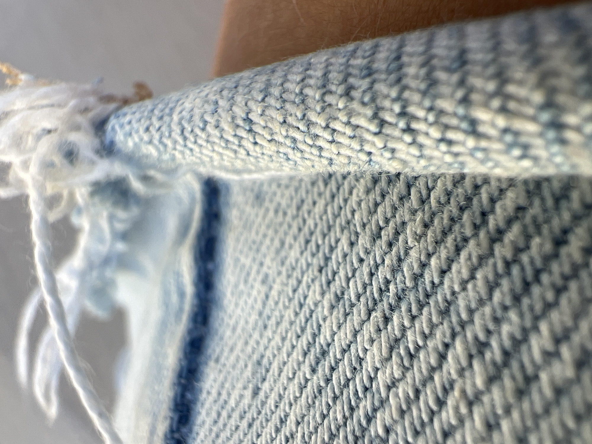 This is a close-up, macro photograph of a rolled-up piece of fabric with a textured weave, likely denim or a similar material, in shades of light blue and white. The fabric is tightly woven, with visible threads forming a diagonal pattern. The edge of the fabric is frayed, with loose white threads creating a soft, fuzzy texture. The fabric is being held by a person's hand, visible on the right side of the image, with skin tone suggesting it is a human hand. The background is out of focus, appearing as a soft, neutral gray. There is no indication of Fontanka, Ukraine, or any urban environment in the image.