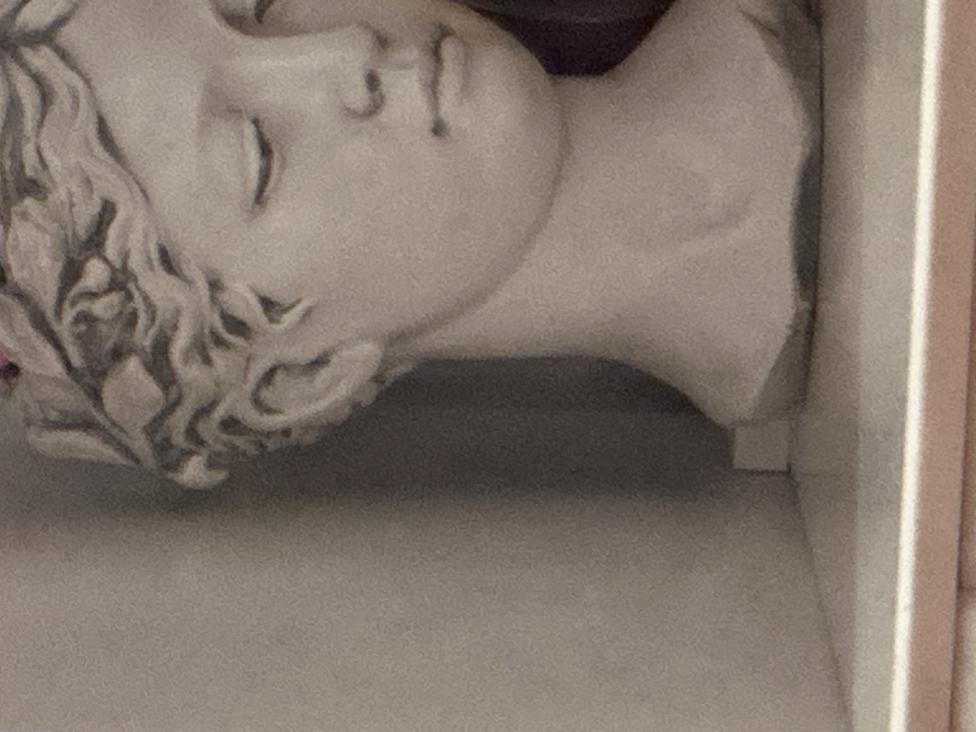 The image displays a close-up, low-angle shot of a plaster bust of a young woman adorned with a laurel wreath, positioned on a shelf against a muted, textured background. The bust is primarily in profile, with its face turned to the right. The lighting casts subtle shadows, highlighting the contours of the facial features, including closed eyes, a straight nose, and slightly parted lips. The texture of the plaster is visible, and the overall color palette is neutral, dominated by shades of beige and gray. The background appears to be a plain wall or partition, creating a simple and unobtrusive setting for the sculpture. The scene is static and devoid of any human presence or activity. The origin or location of this bust, whether in Georgiyevsk, Russia, or elsewhere, cannot be determined from the visual information provided.
