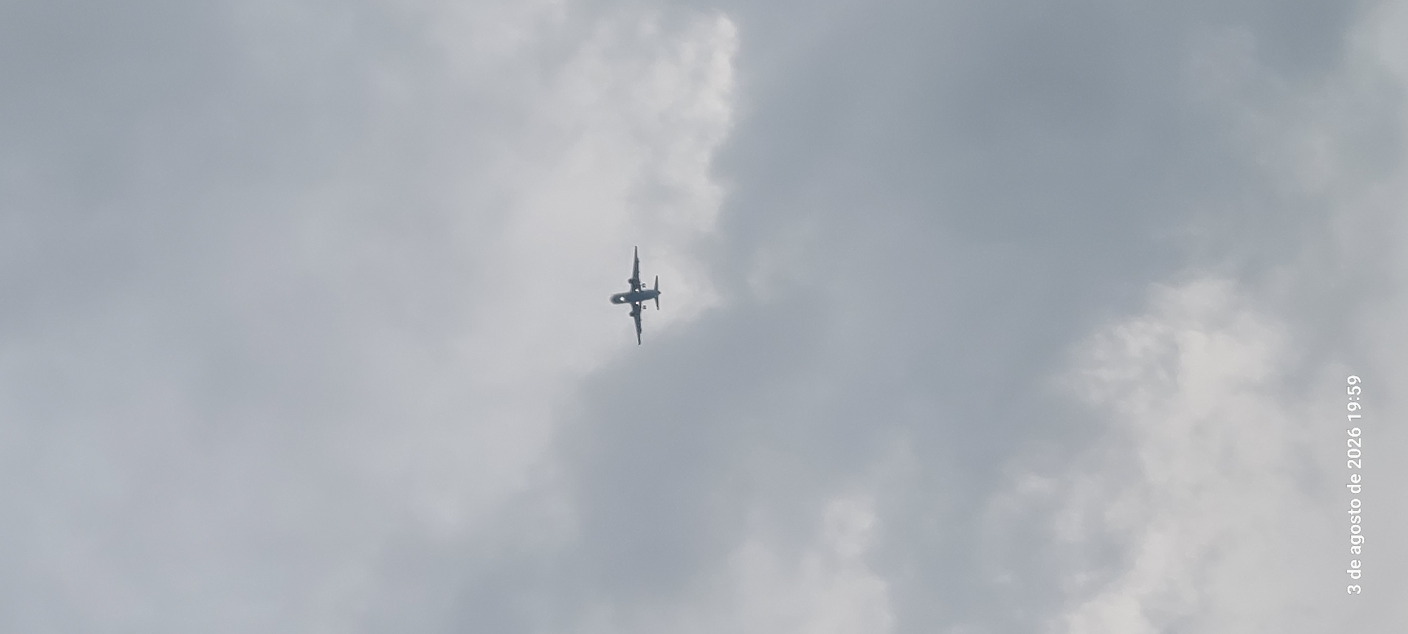 A lone airplane is captured in flight against a cloudy, overcast sky. The aircraft, silhouetted against the muted light, appears to be a commercial passenger jet, possibly on approach or departure. Its landing gear is extended, suggesting it is either landing or preparing for takeoff. The plane is positioned in the center of the frame, against a backdrop of diffused gray clouds, creating a stark, solitary image. The timestamp at the bottom of the image indicates the photo was taken on August 3, 2026, at 19:59. The image's metadata specifies the location as Castelldefels, Spain, a coastal city known for its proximity to Barcelona and its airport, which would explain the presence of a visible airplane. There are no people or other significant objects or actions depicted in the scene.