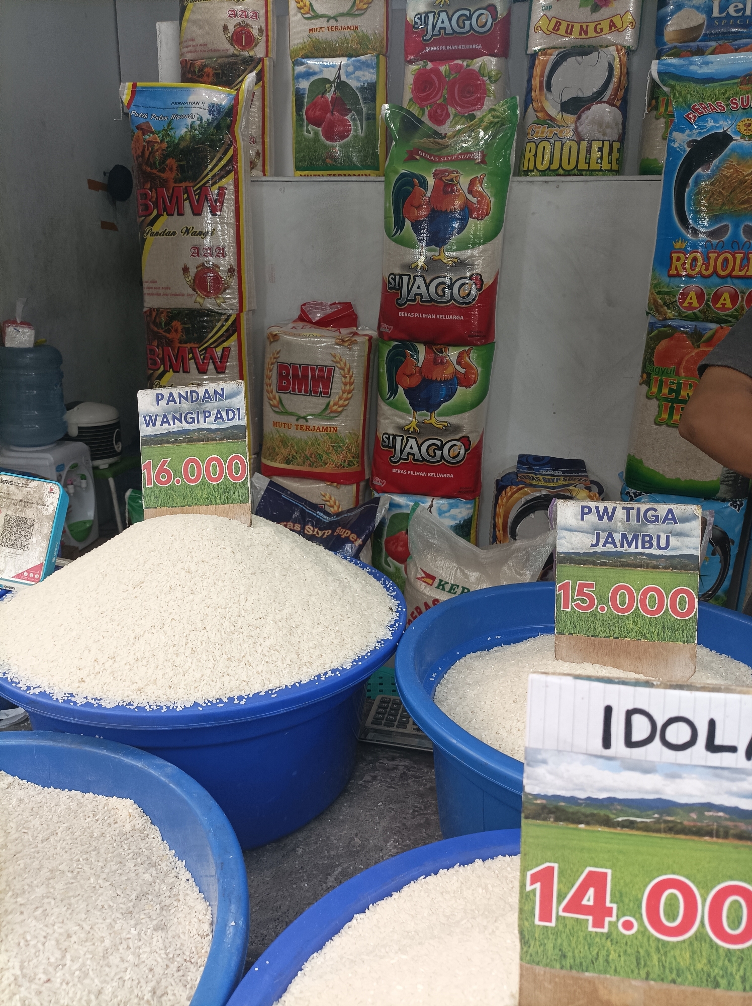 In a retail setting, likely a market stall in West Jakarta, Indonesia, a variety of rice packages are displayed. In the foreground, two large blue plastic tubs are filled with white rice. A price sign for "Pandan Wangi Padi" priced at 16.000 is positioned in front of the larger tub. To the right, another price sign for "PW Tiga Jambu" priced at 15.000 is visible, with a portion of a tub of rice behind it. A third price sign, partially obscured, reads "IDOLA" with the price 14.00. The background features stacked bags of rice, each with distinct branding and imagery, including roosters, cherries, and agricultural scenes. To the left, a water dispenser and electronic devices are present. The overall scene depicts a bustling rice sales point.