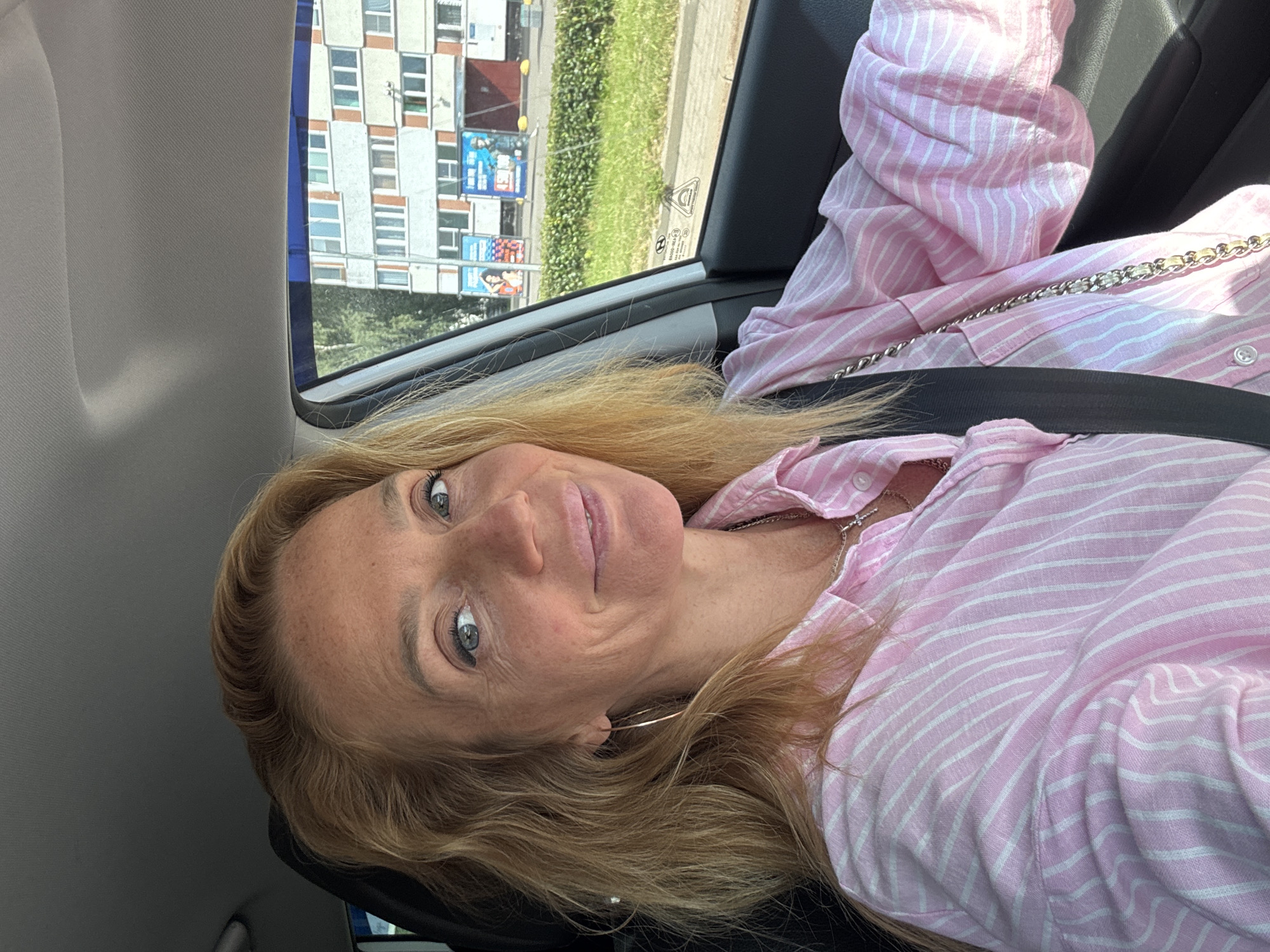 A woman with blonde hair is sitting in the driver's seat of a car. She is wearing a pink and white striped button-down shirt and a silver necklace. Her seatbelt is fastened. Through the car window, a residential building with multiple floors and windows can be seen. The building appears to be in an urban setting, with trees and grassy areas visible in the foreground and background. The scene is illuminated by daylight. The location is identified as Sinyavino, Russia.