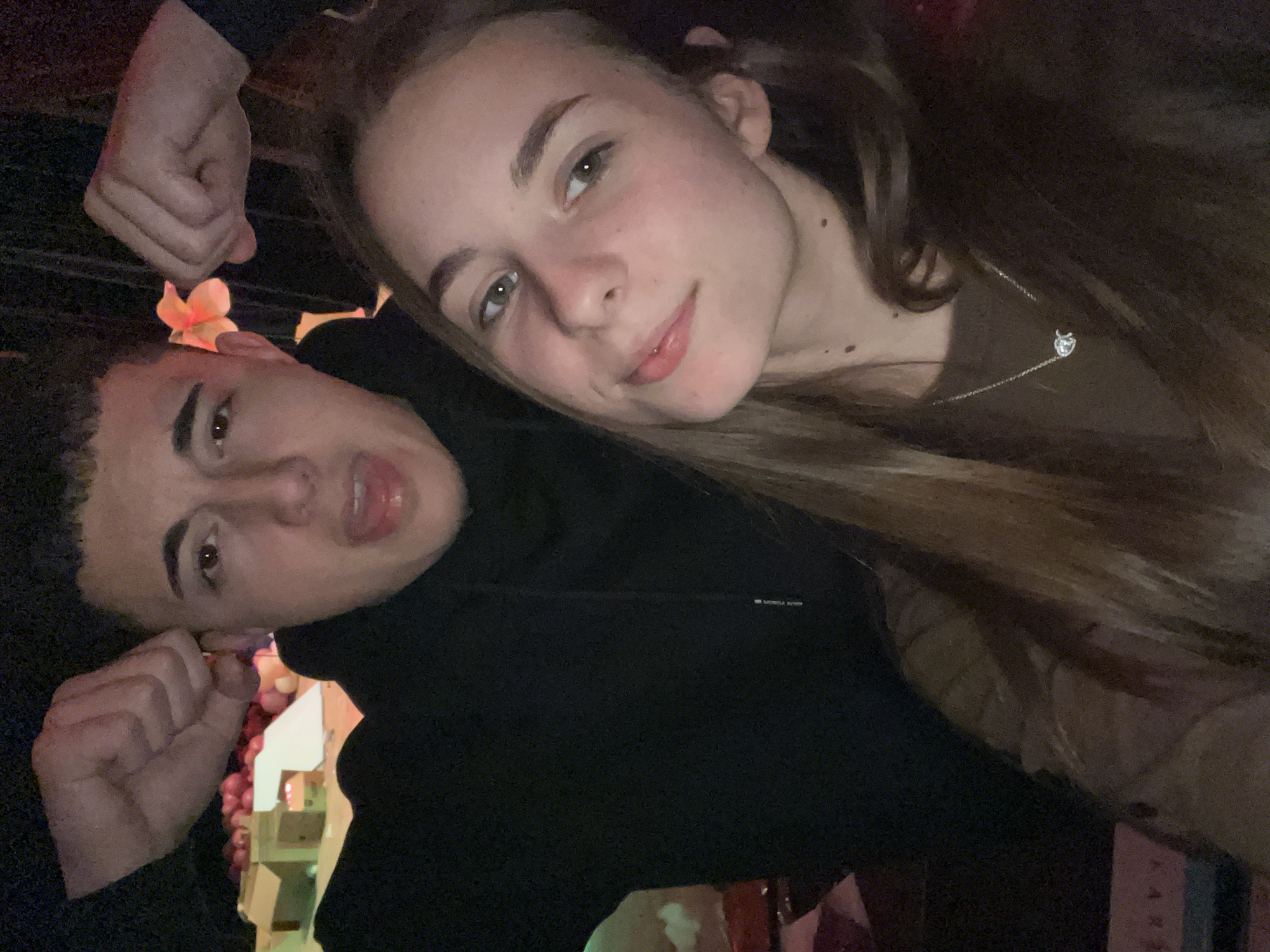 A close-up, slightly angled, selfie-style photograph shows a young man and woman. The woman, positioned above and to the right, leans her head towards the man. She has fair skin, light eyes, and medium-length brown hair, and wears a delicate silver necklace. The man, below and to the left, has short dark hair and appears to be making a playful expression. He wears a dark-colored hoodie. Two fists are raised near the sides of their faces, possibly belonging to a third person, as one fist has a small flower on it. In the blurred background, one can discern some decorative elements, including what appear to be artificial grapes and decorative structures. A neon sign with the word “KARA” is visible in the lower right corner. There is no information given to indicate the location is in Nova Zagora, Bulgaria.