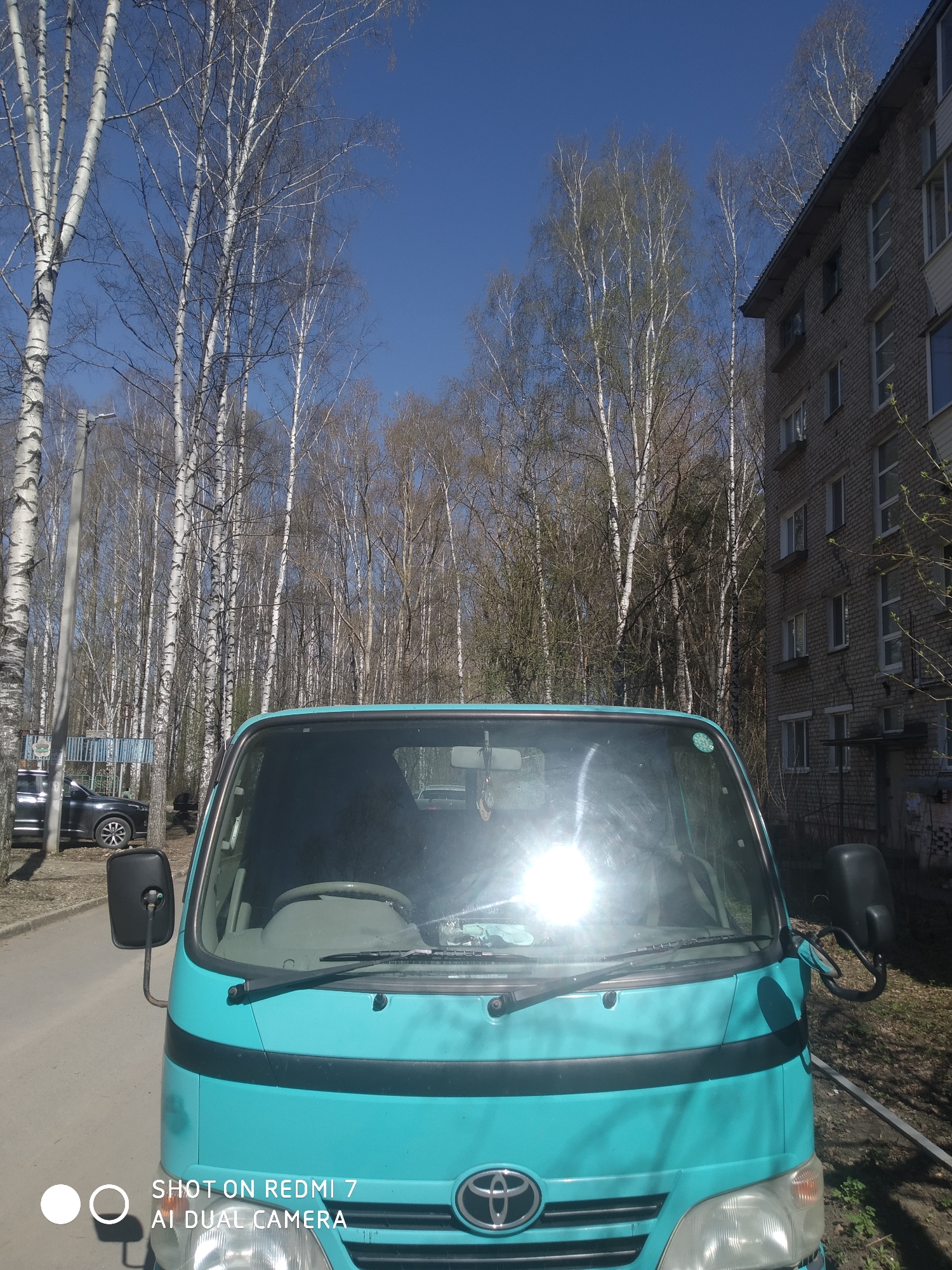 The image depicts a turquoise Toyota truck parked on a street in Chaikovsky, Russia. The truck is positioned in the foreground, with its windshield reflecting the sun. Behind the truck are birch trees lining the street. To the right of the truck is a grey brick building. A black car is parked further down the street on the left. The sky is clear and blue. The camera's timestamp is visible at the bottom of the image.