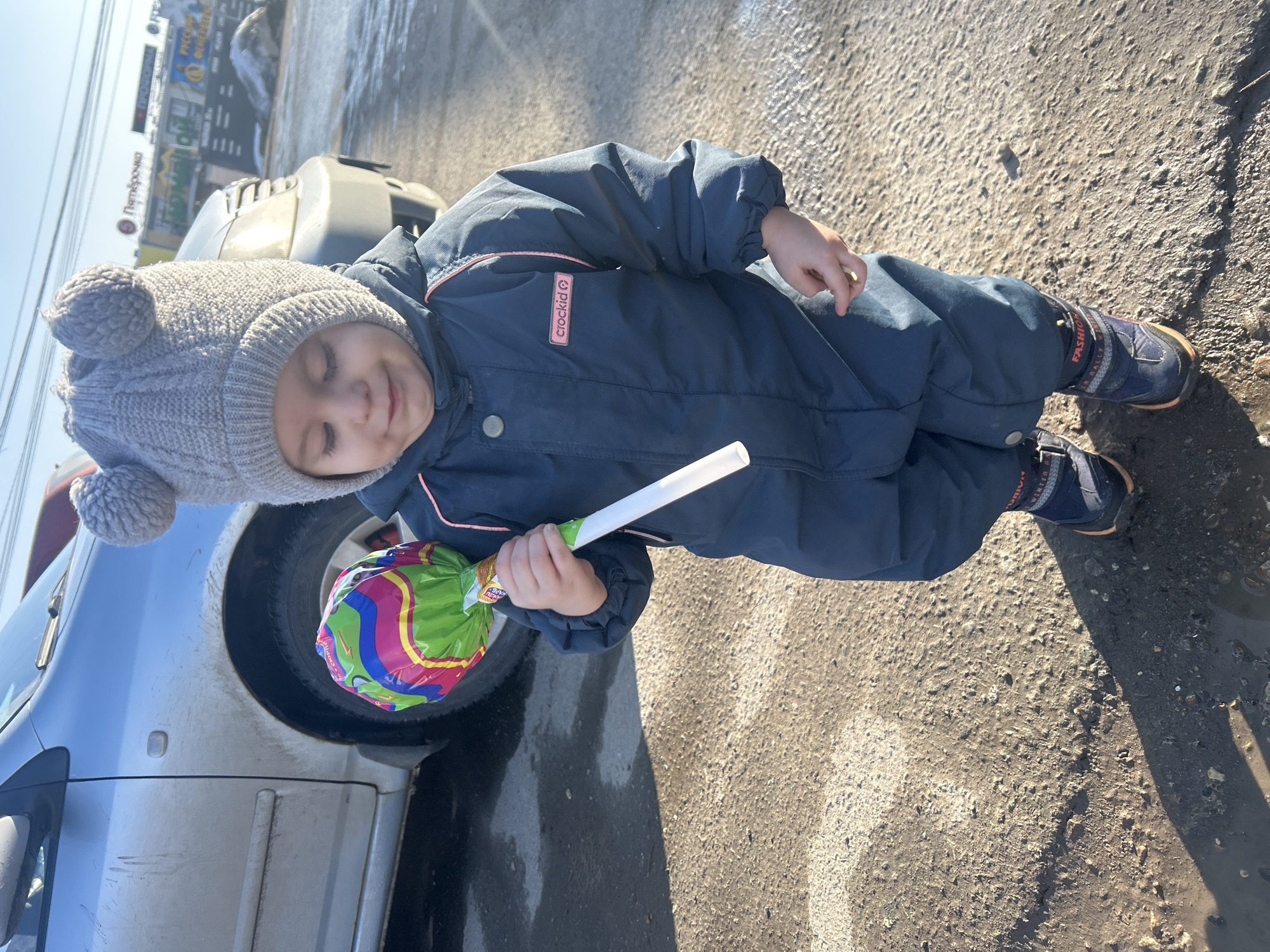 A young child, appearing to be a toddler, stands on a rough, damp asphalt surface with visible dirt and minor puddles, casting a long shadow to their left under bright sunlight. The child wears a grey knitted hat with two pom-pom-like ears, a navy blue or dark teal full-body "crockid" brand winter suit with pink piping, and dark blue boots with orange accents. They are looking downwards with a soft expression, holding a large, brightly colored lollipop in their right hand. The lollipop is wrapped in clear plastic featuring wavy patterns of green, pink, blue, and yellow, with a white stick. In the background to the left, a dirty silver sedan is partially visible. Further back, across a parking area, commercial buildings are visible under a clear blue sky. A prominent red sign on one building displays "Пятёрочка" (Pyaterochka) in white Cyrillic text. Below it, other signs with Cyrillic text include "РУССКИЙ ДЕЛИКАТЕС" (Russian Delicatessen) and "СПОРТ" (Sport). Patches of melting snow or ice are present in the distant background. The scene is located in Chaikovsky, Russia.