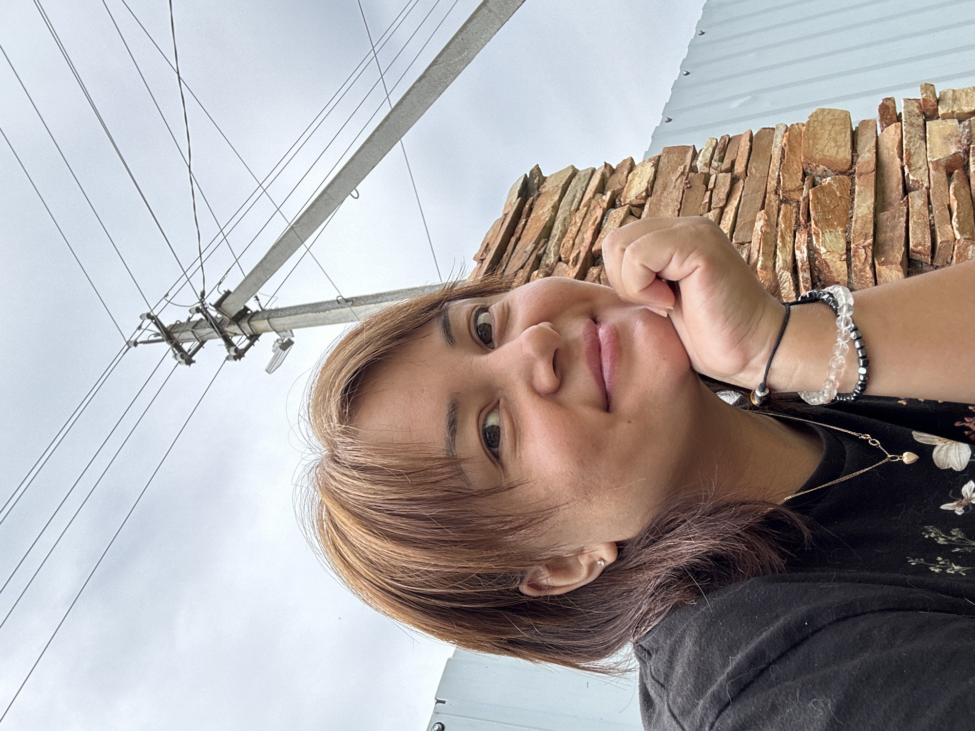 A woman is captured in a close-up, outdoor selfie. She has shoulder-length, auburn hair and is looking towards the camera with a gentle smile, her right hand resting on her chin. She is wearing a black t-shirt with a delicate gold necklace featuring a small heart pendant. On her left wrist, she wears two bracelets, one with clear beads and another with dark, metallic beads.
The background features a grey, overcast sky with visible power lines and a utility pole. To the right of the woman, a textured wall made of stacked, reddish-brown stones forms a visual element. Behind the stone wall, a light blue, corrugated metal fence is visible. The overall setting appears to be an outdoor space, possibly a backyard or a less urbanized area, given the presence of utility infrastructure and rustic fencing. The lighting suggests it is daytime, but overcast conditions have diffused the light.