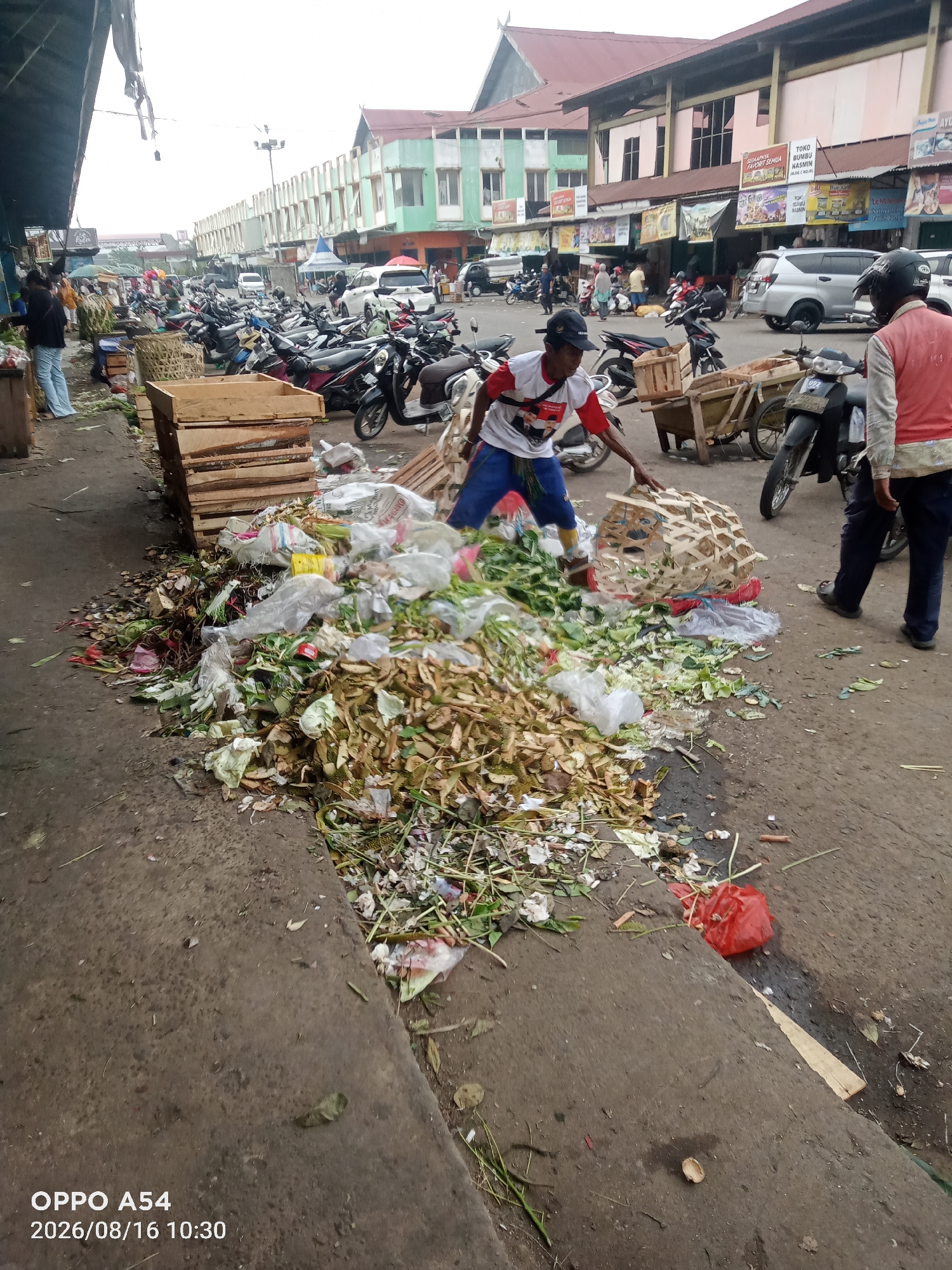 The image depicts an outdoor marketplace scene in Jambi, Indonesia, possibly at a traditional market. A large pile of organic waste, consisting of vegetable scraps, durian rinds, and other food waste, is accumulated on the ground in the foreground. Two men are present. One man, wearing a red vest over a white shirt and blue pants, is standing to the right, partially visible. The other man, in a white t-shirt with red accents and blue pants, is actively working with a large, woven basket, seemingly engaged in collecting or moving the waste. The background shows several rows of parked motorcycles, various market stalls with awnings and signage, and buildings that appear to be part of the market complex. The environment appears to be overcast, and the ground is paved with concrete and asphalt, with some areas wet and stained. The overall impression is a busy, working marketplace with sanitation challenges.