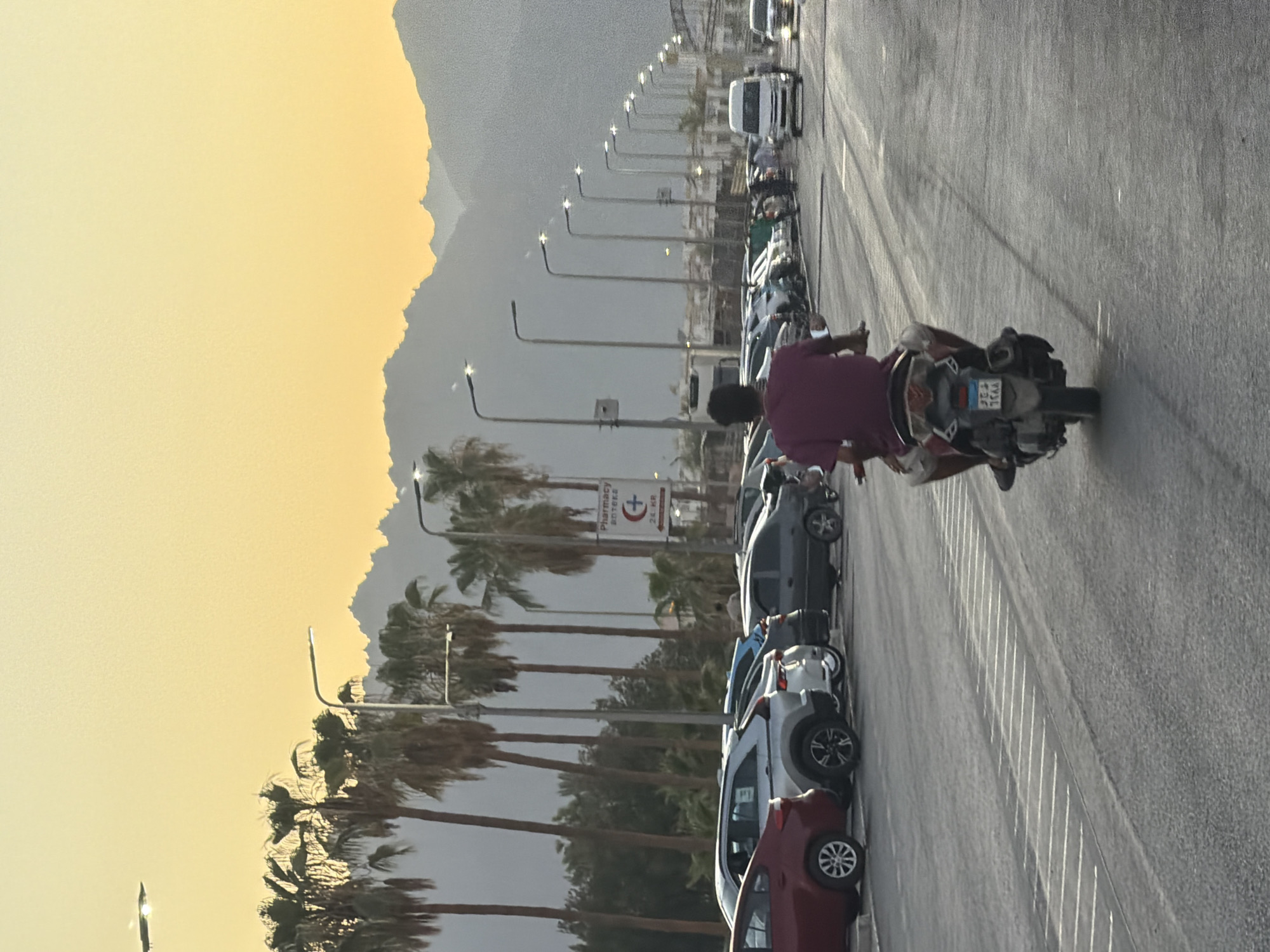 A person rides a motorcycle on a street lined with palm trees and parked cars, with mountains in the background under a yellow sky, likely during sunset. A pharmacy sign is visible on the right. The scene appears to be in Sharm El Sheikh, Egypt, a popular tourist destination known for its coastal location and desert landscape.