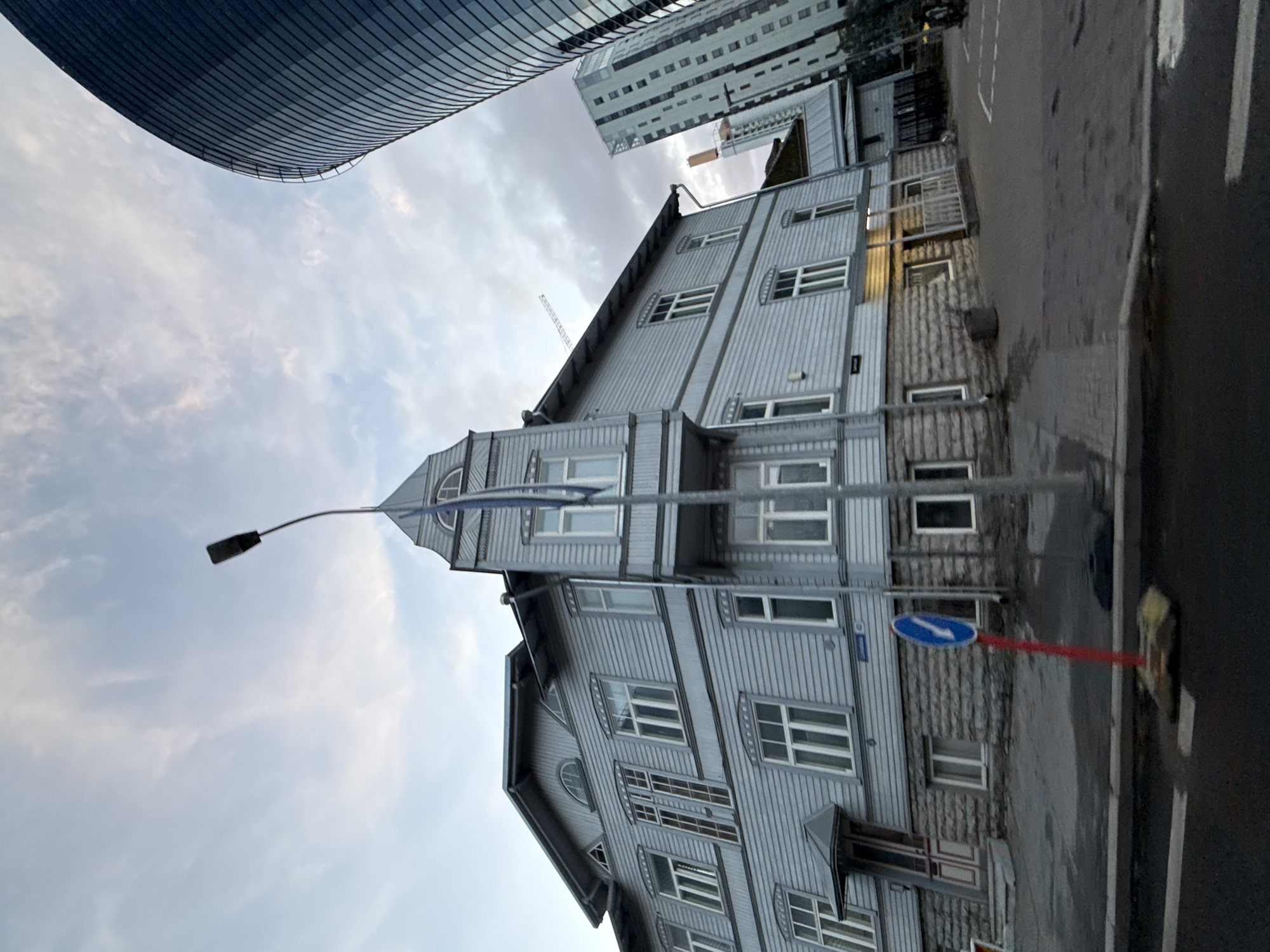 The image captures a street scene in Tallinn, Estonia, featuring a traditional light-colored wooden house with a distinctive tower structure. Adjacent to it, a modern glass-fronted skyscraper curves upwards, contrasting sharply with the historical architecture. A taller, more rectangular, white skyscraper is also visible in the background. The sky above is partly cloudy. In the foreground, a blue and white directional sign on a red pole stands on the pavement. The street appears wet, suggesting recent rain.