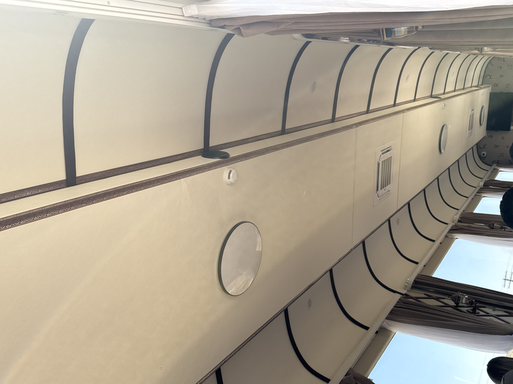 The image shows the interior ceiling of what appears to be a train carriage, with beige panels and dark decorative trim. There are several circular light fixtures, a smoke detector, and an air conditioning unit visible on the ceiling. Curtains are visible along the side of the carriage, and through the windows, a bright sky and some distant power lines can be seen. The scene is indoors, likely on a mode of transportation. No specific location in the city of Kok-Moinok - 1, Kyrgyzstan can be determined from the image alone. There are no discernible people or actions in this image.