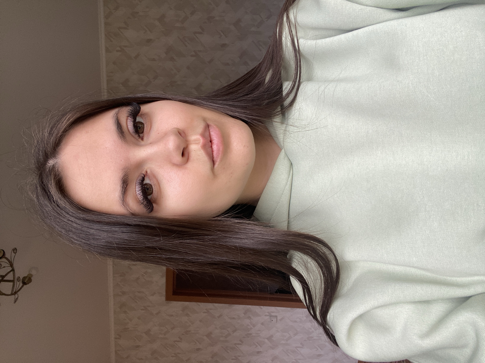A woman with long brown hair and false eyelashes is seen indoors, likely in a residential setting. She wears a light green sweatshirt. Behind her is a wall with patterned wallpaper. A framed object is partially visible on the wall, along with a decorative light fixture.
The location, Zhigalki, Russia, cannot be confirmed from visual information.