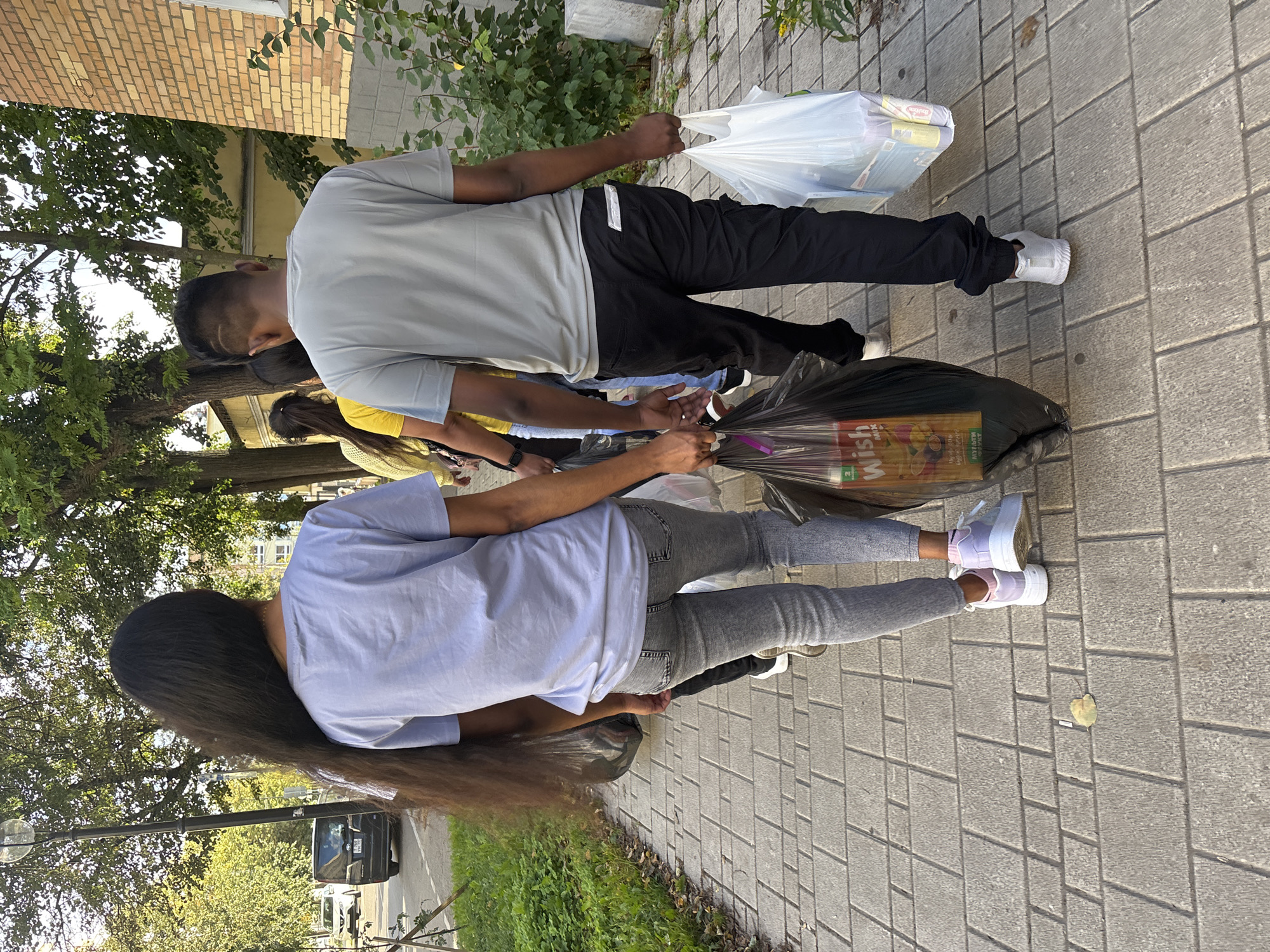 The image depicts a group of young people walking on a sidewalk in the city of Zvenigorod, Russia. The foreground is dominated by the backs of three individuals. The person on the left is a female with extremely long, dark hair, wearing a light blue t-shirt and grey jeans. She is carrying a black plastic bag in her right hand, which contains what appear to be food items, including a box labeled "Wish Mix". The person in the center is a male wearing a grey t-shirt and black cargo pants. He is carrying a large white plastic bag in his right hand, filled with packaged goods, possibly groceries. To the right of him, partially visible, is another individual. The sidewalk is paved with grey rectangular bricks.
In the background, other people can be seen walking ahead. To the left of the sidewalk, there is a grassy area with some foliage and trees. Further in the background, a dark-colored car is parked on the street, and buildings with brick facades are visible. The scene appears to be set on a sunny day, with trees lining the street and providing shade. The overall impression is of a casual outing or returning from shopping.