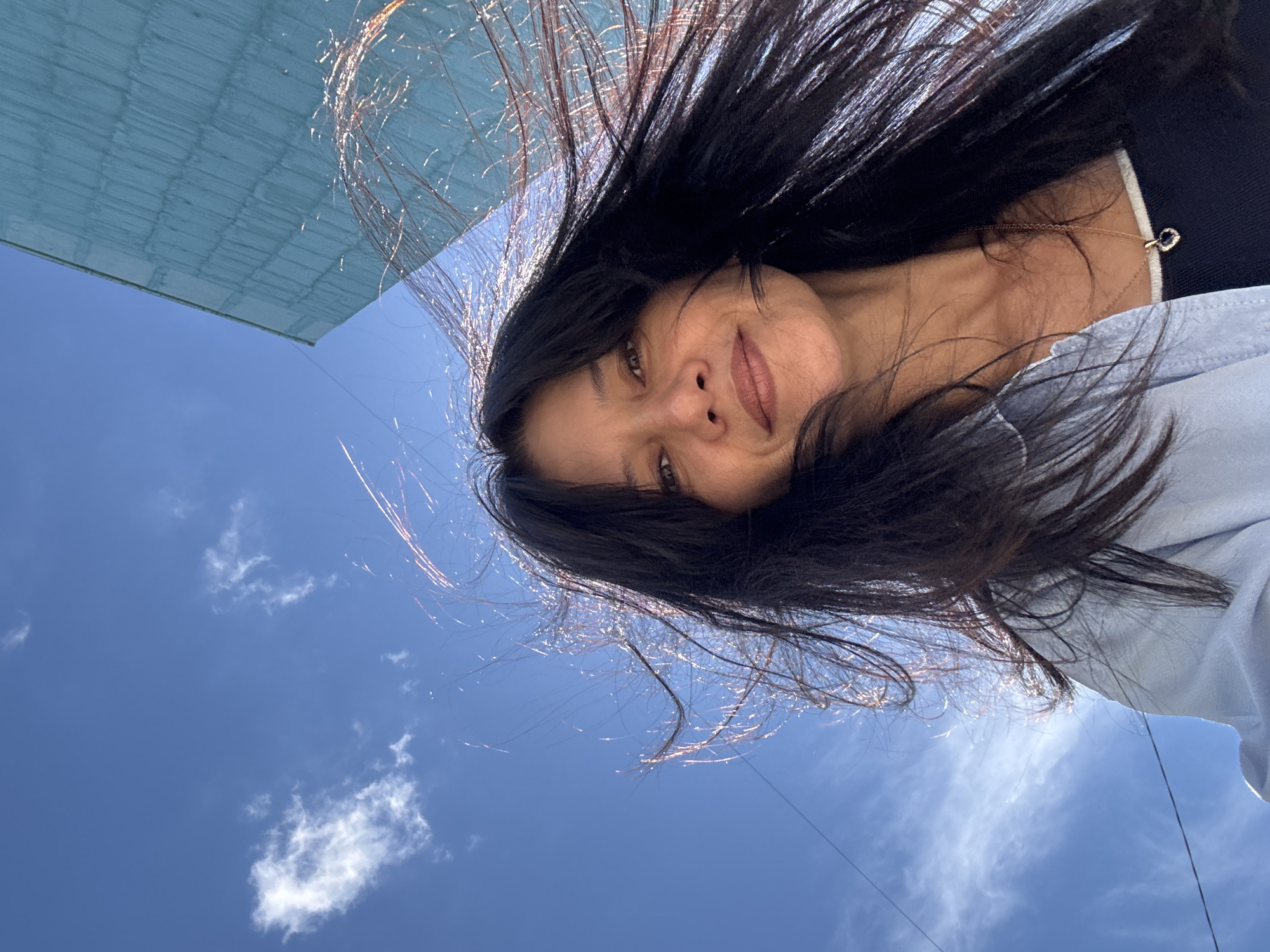 A woman is captured in a low-angle selfie against a bright blue sky with scattered white clouds. Her long, dark hair is windswept, with strands catching the sunlight and appearing almost metallic. She is wearing a light blue collared shirt over a dark top, and a delicate gold necklace with a small heart-shaped pendant. The background features a corner of a teal-colored building with a textured surface, possibly brick or stucco, and a thin black wire, likely a power or telephone line, stretches across the sky. The overall impression is of an outdoor, possibly urban environment on a sunny day. The specific location in Chaikovsky, Russia, is not discernible from the visual information in the image.