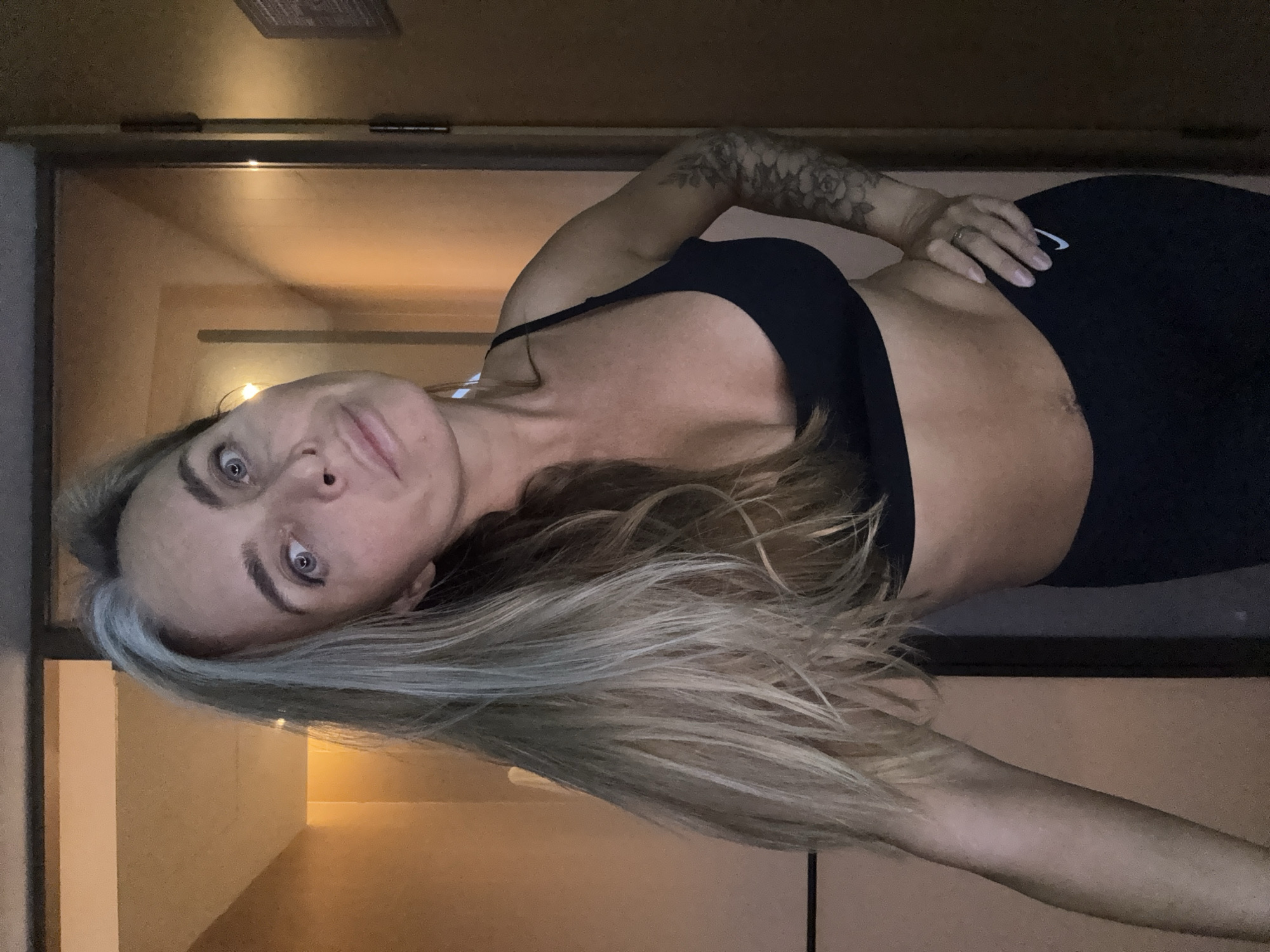 In this medium shot, a woman with long, blonde hair, some of which is gray, stands in what appears to be a hallway or corridor. She is looking directly at the camera with her head tilted. She is wearing a black sports bra and black leggings. Her left arm is bent at the elbow, with her hand resting on her hip. She has a tattoo on her right arm. The hallway is dimly lit, with warm-toned walls and an open door visible on the right side of the frame. The location is not specified as Shanghai, China.