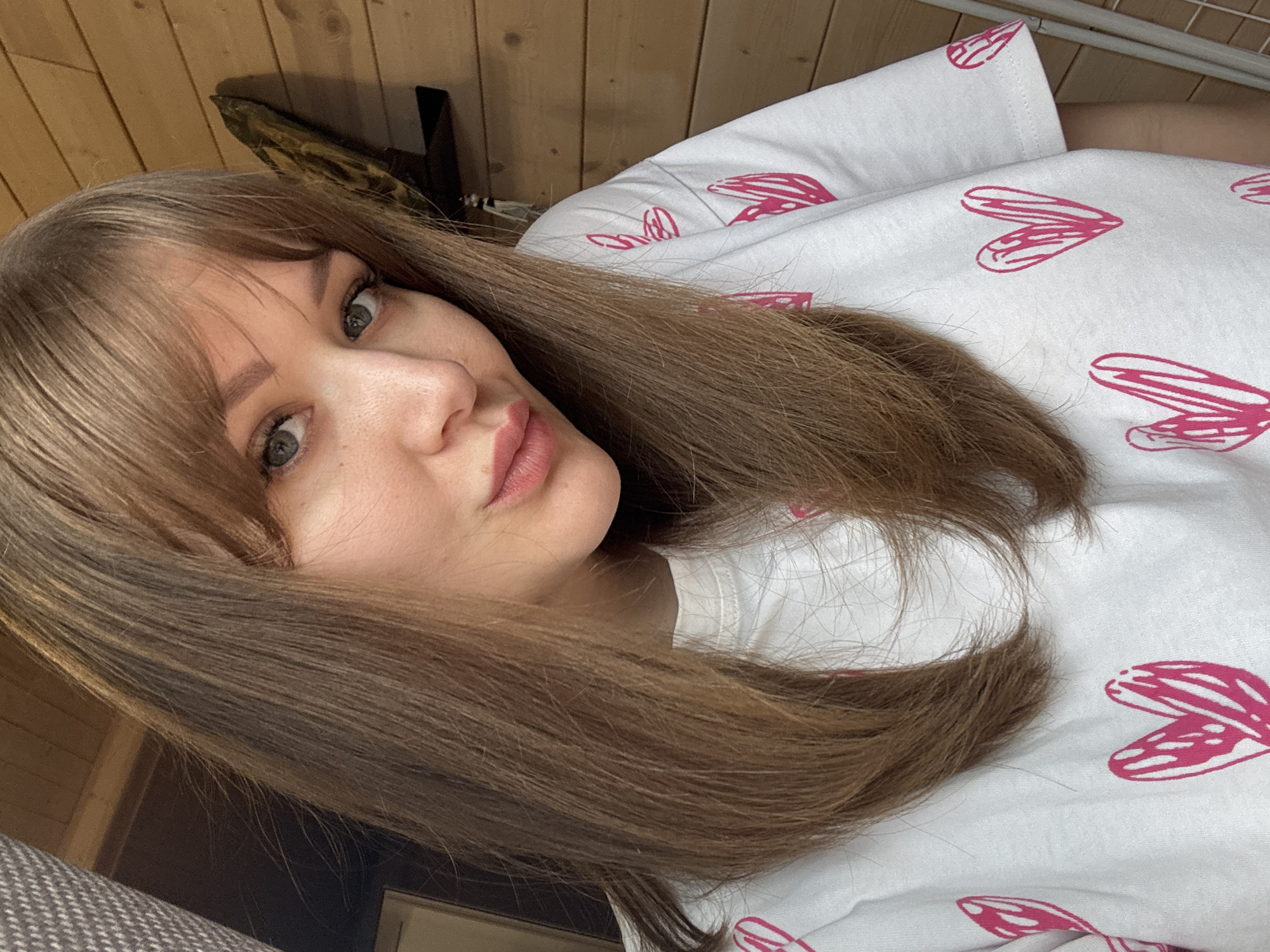 A woman with long, light brown hair and bangs is looking at the camera. She is wearing a white t-shirt with pink heart designs. The background is a wood-paneled wall. The location is likely indoors and no information or indication the image was taken in Taman, Russia.