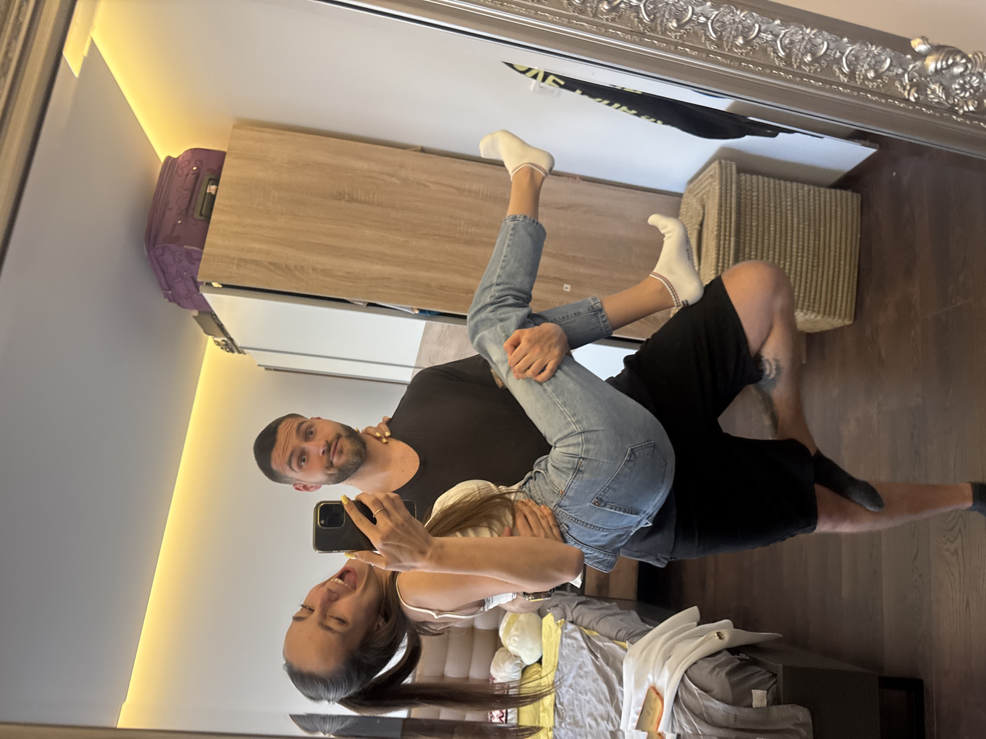 A couple in Sofia, Bulgaria, is reflected in a large ornate mirror. A woman holds a cell phone, taking a photo of the reflection. The man is seated, wearing black shorts and a black T-shirt; the woman is straddling him in light-colored jeans, a white top, and white socks. A gray suitcase and wooden wardrobe cabinet are in the background. A woven hamper, bed, and bedding are also visible.