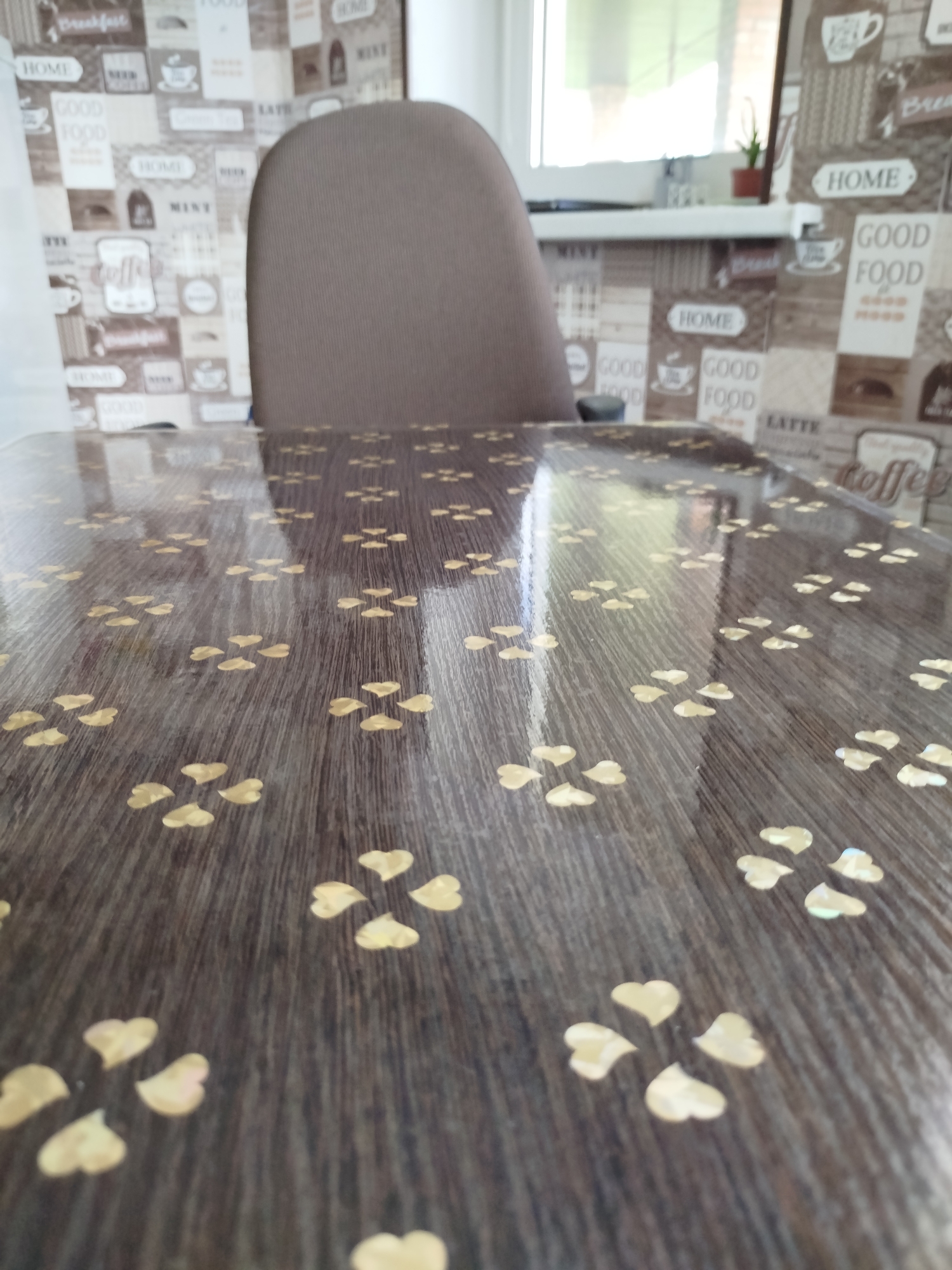 The image shows a close-up of a dark wood-patterned table with a glossy finish. Scattered across the table are numerous small, gold, heart-shaped sequins, arranged in clusters resembling four-leaf clovers. The background is blurred but appears to be a wall with a patterned wallpaper featuring text like "GOOD FOOD" and coffee cup graphics. A portion of a brown chair is visible behind the table. The lighting suggests an indoor environment, possibly a dining area or kitchen. The scene is static and does not depict any people or actions. No specific location in Крывасельскі сельскі Савет, Belarus can be determined from the visual information alone.