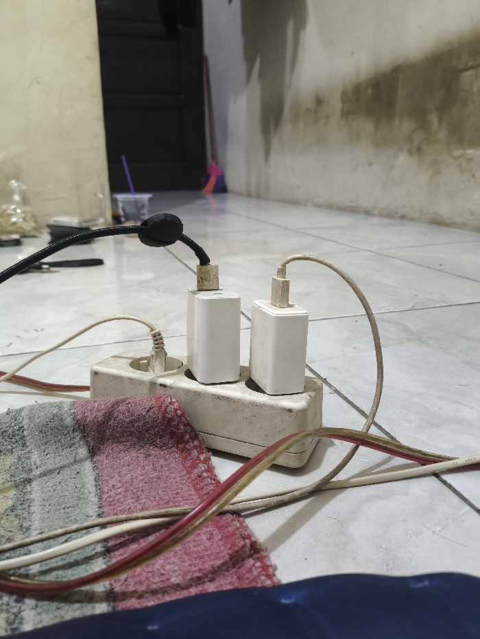 The image presents a low-angle, close-up view of a dirty white power strip on a light-colored tiled floor, in what appears to be an indoor setting in Cibatu, Indonesia. Three charger adapters are plugged into the power strip. Two prominent white rectangular phone chargers are visible, with their cables (one black, one white) extending upwards. Another cable is plugged into the leftmost socket. The power strip itself is heavily soiled and discolored.
In the immediate foreground, a piece of striped fabric, featuring red, green, and white colors, lies partially visible next to the power strip, along with a dark blue object that could be a bag or another piece of cloth. Multiple charging cables, including distinct red and brown ones with faint, indistinct white text (possibly including "401 W"), are tangled around the power strip and on the floor.
The background shows a simple, somewhat unkempt room. A light-colored wall on the right is heavily stained with dark, irregular marks, suggesting dampness or age. To the left, a dark doorway or door is partially visible, next to another light-colored wall. Further in the background, a purple straw protrudes from a clear cup or bottle, and the handle of a broom (pink/purple and yellow/orange) leans against a wall.
The scene depicts an informal charging area, possibly in a utility space or a general living area, with an emphasis on functional objects. No people are present. The overall impression is one of everyday use in a modest, well-worn environment.