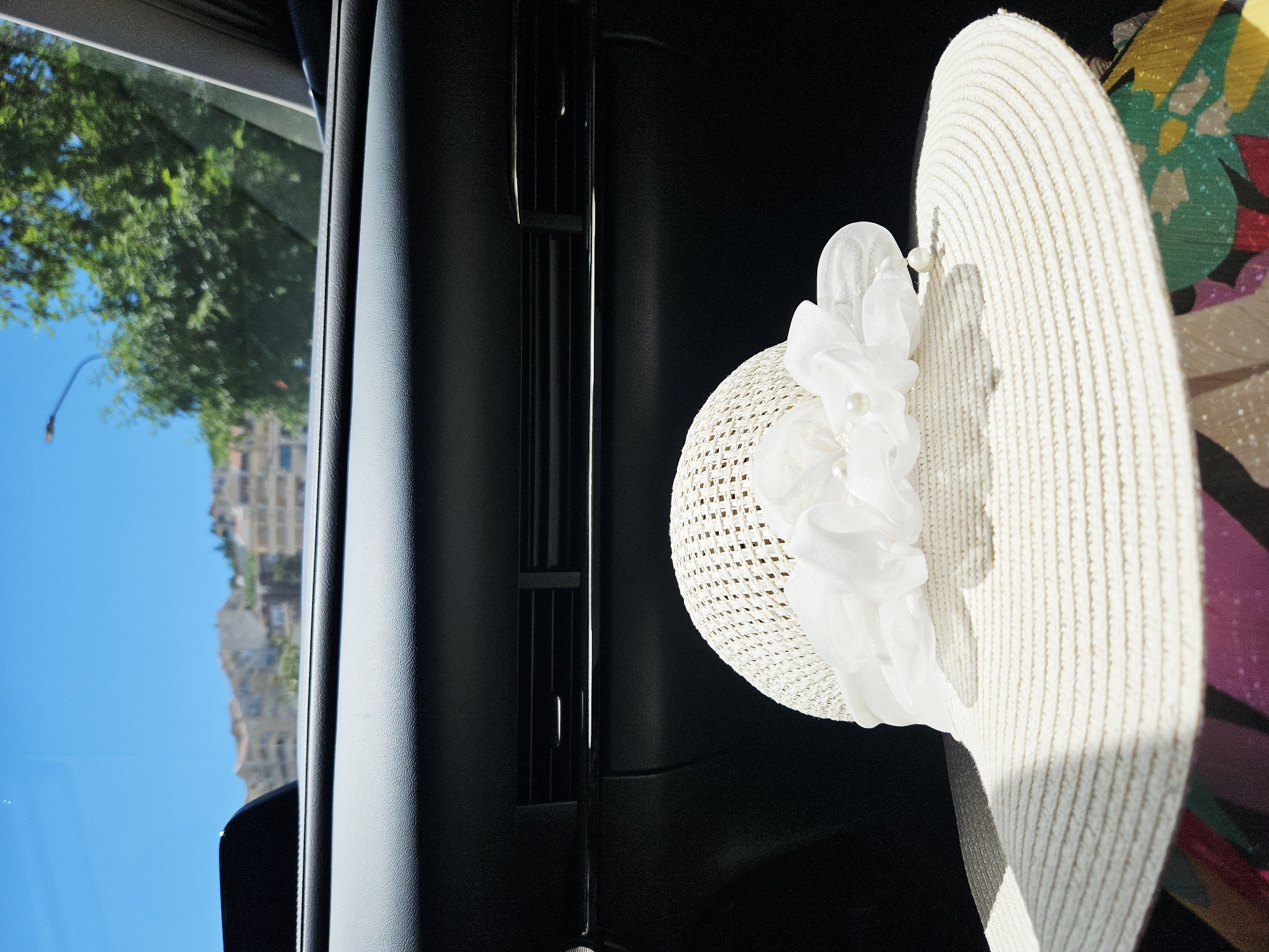 The image shows the interior of a car. Outside the window, one can see a building, blue sky, and trees. Attached to the dashboard is a white hat decorated with a white flower. Part of a colorful wall is also visible. The location is Unknown, Unknown.