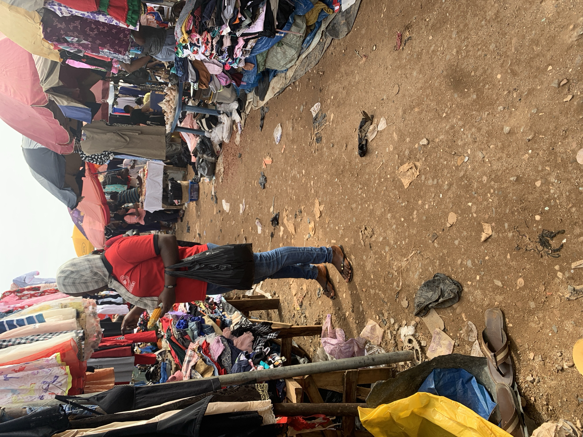 This is a photograph of an outdoor market in Nyanya, Nigeria. The ground is dirt and covered with debris, including plastic bags, paper, and other refuse. In the foreground, a person wearing a red shirt and blue jeans is standing in front of a stall displaying various clothing items. The person is holding a cob of corn and carrying a black plastic bag. The market appears to be selling used clothing, as indicated by the piles of garments on display and on the ground. Several other people can be seen in the background, as well as market stalls and umbrellas providing shade. The overall scene depicts a bustling, somewhat disorganized, marketplace environment.