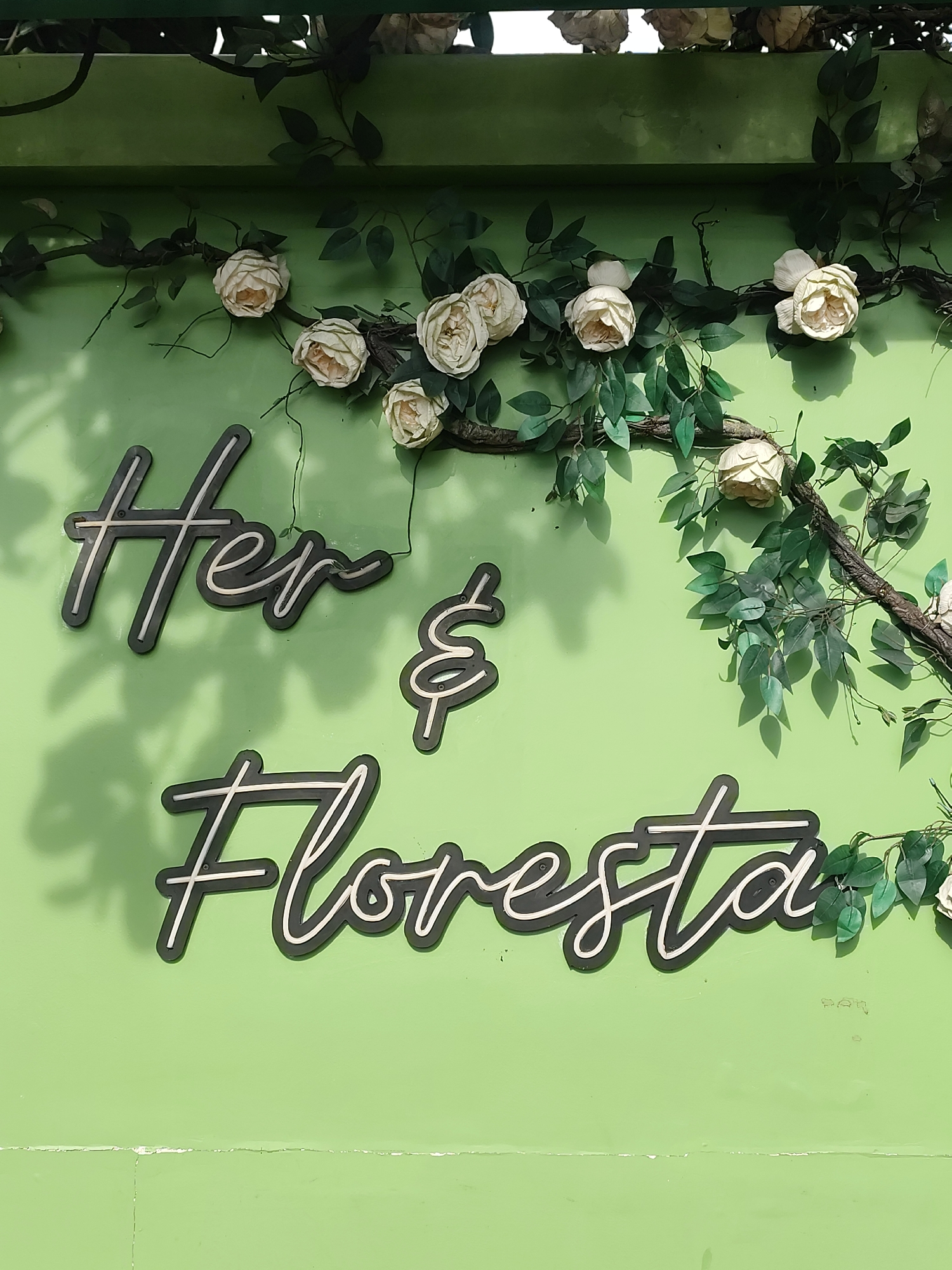The image shows a neon sign on a light green wall. The sign reads "Her & Floresta" in stylized script lettering. The sign is dark with white neon accents. There are artificial flowers and green vines draped over and around the sign. The overall aesthetic is romantic and natural, with the light green wall providing a vibrant backdrop. The scene is set outdoors, possibly on a facade or a dedicated display wall in a public space. The lighting suggests it is daytime, with shadows cast by the sign and foliage onto the wall. The specific location in Da Lat, Vietnam is not discernible from the image itself, as there are no identifying landmarks or cultural markers present.