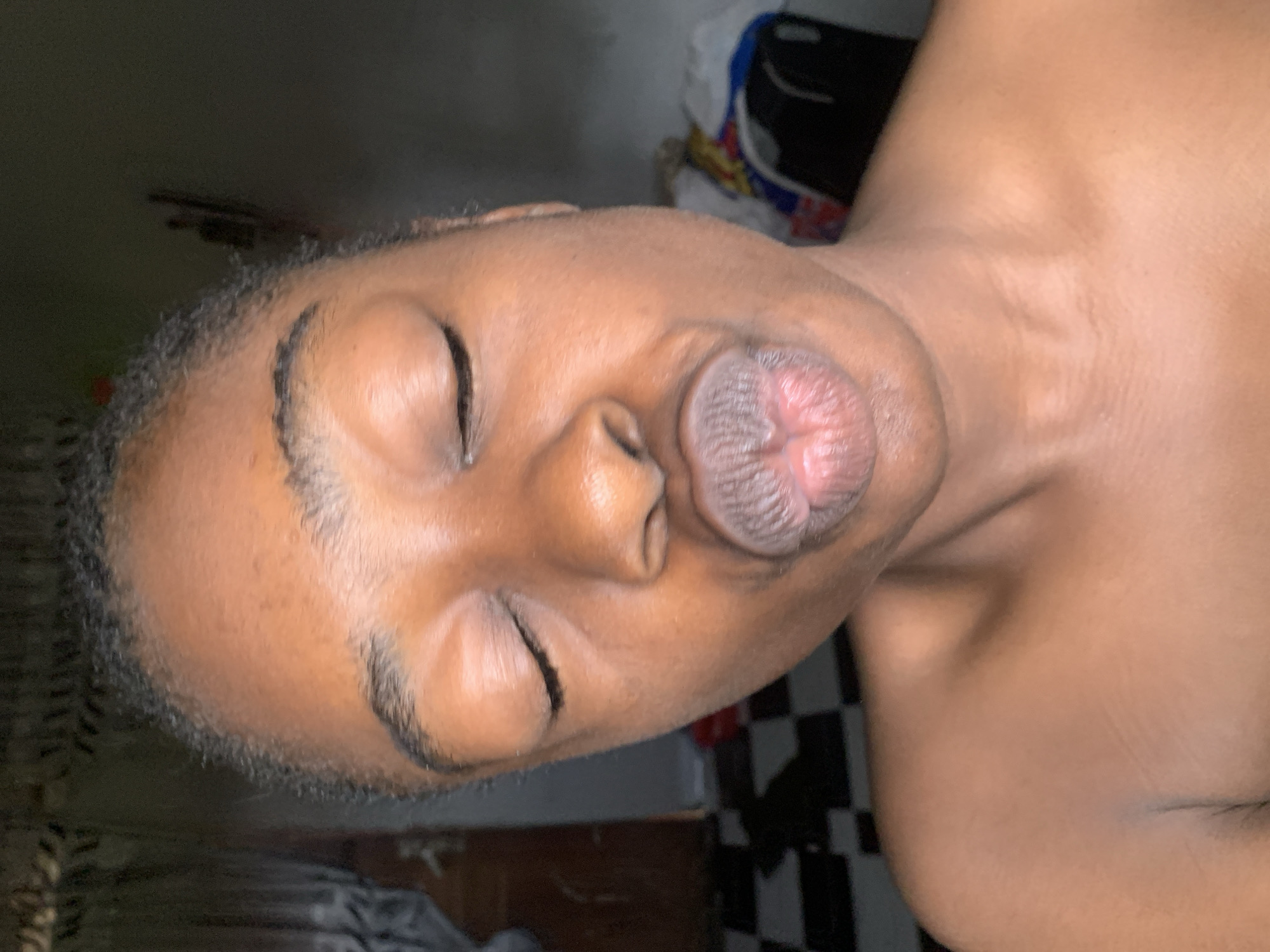 A medium close-up, portrait-style photograph shows a Black individual with their eyes closed and lips pursed, as if blowing a kiss. The individual's head is slightly tilted to the left, and their shoulders and upper chest are visible. The skin tone is a warm brown. The background is dimly lit and appears to be an indoor setting, with a tiled floor and some indistinct objects, possibly furniture and clothing, visible behind the subject. There is no indication of the specific location within Rafin Par, Nigeria, other than it being an indoor environment.