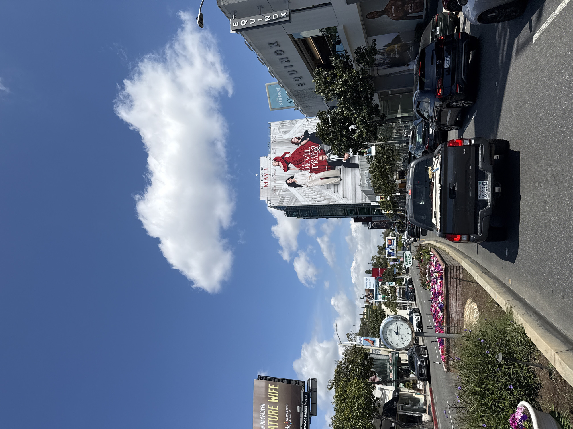 The image captures a street scene in West Hollywood, USA. Prominently featured is a large billboard advertising the movie "The Devil Wears Prada," with the release date of May 1. A smaller billboard promotes the show "Signature Wife." Buildings visible include structures with the words "Equinox" and "Sunset" on them. The scene is set on a street with multiple cars and a street clock. There are trees and flower beds along the side of the road. The sky is blue with scattered clouds.