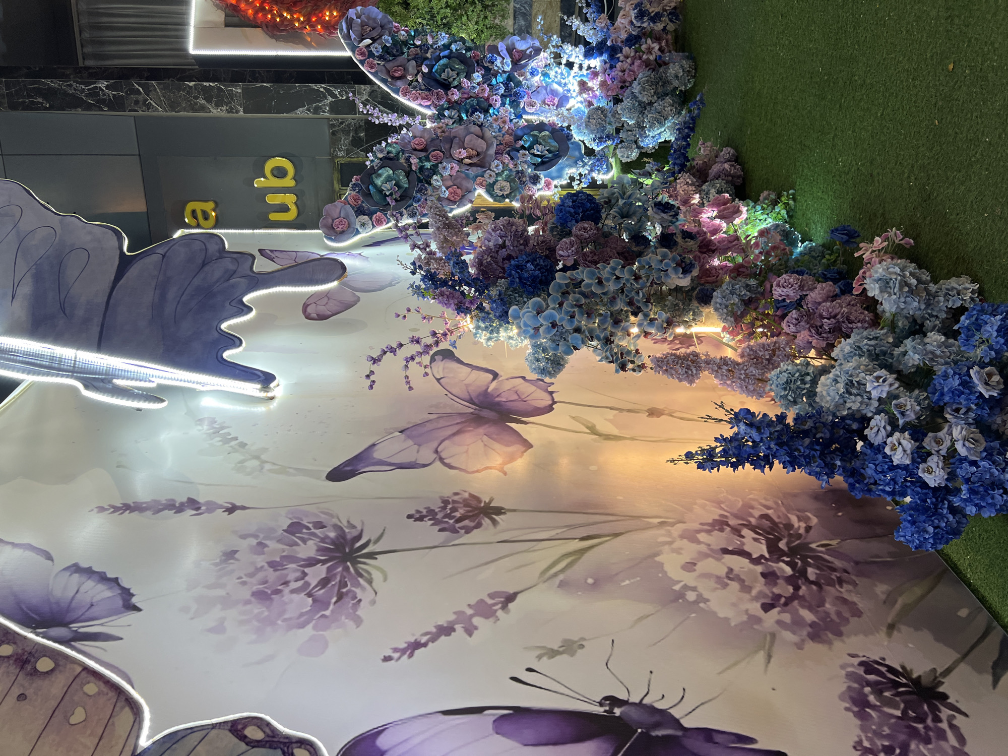 The image depicts an outdoor decorative display featuring large purple butterfly wing cutouts against a wall mural of purple flowers and butterflies. The display is adorned with abundant artificial flowers in shades of blue, purple, and pink, some of which are illuminated by string lights. The ground is covered with artificial green grass. In the background, a building facade with dark marble accents and yellow signage spelling out "a ub" is visible, suggesting a commercial establishment. The overall scene is festive and likely intended for decorative purposes, possibly at an event or a commercial venue in Nha Trang, Vietnam. No people are present in the frame.
