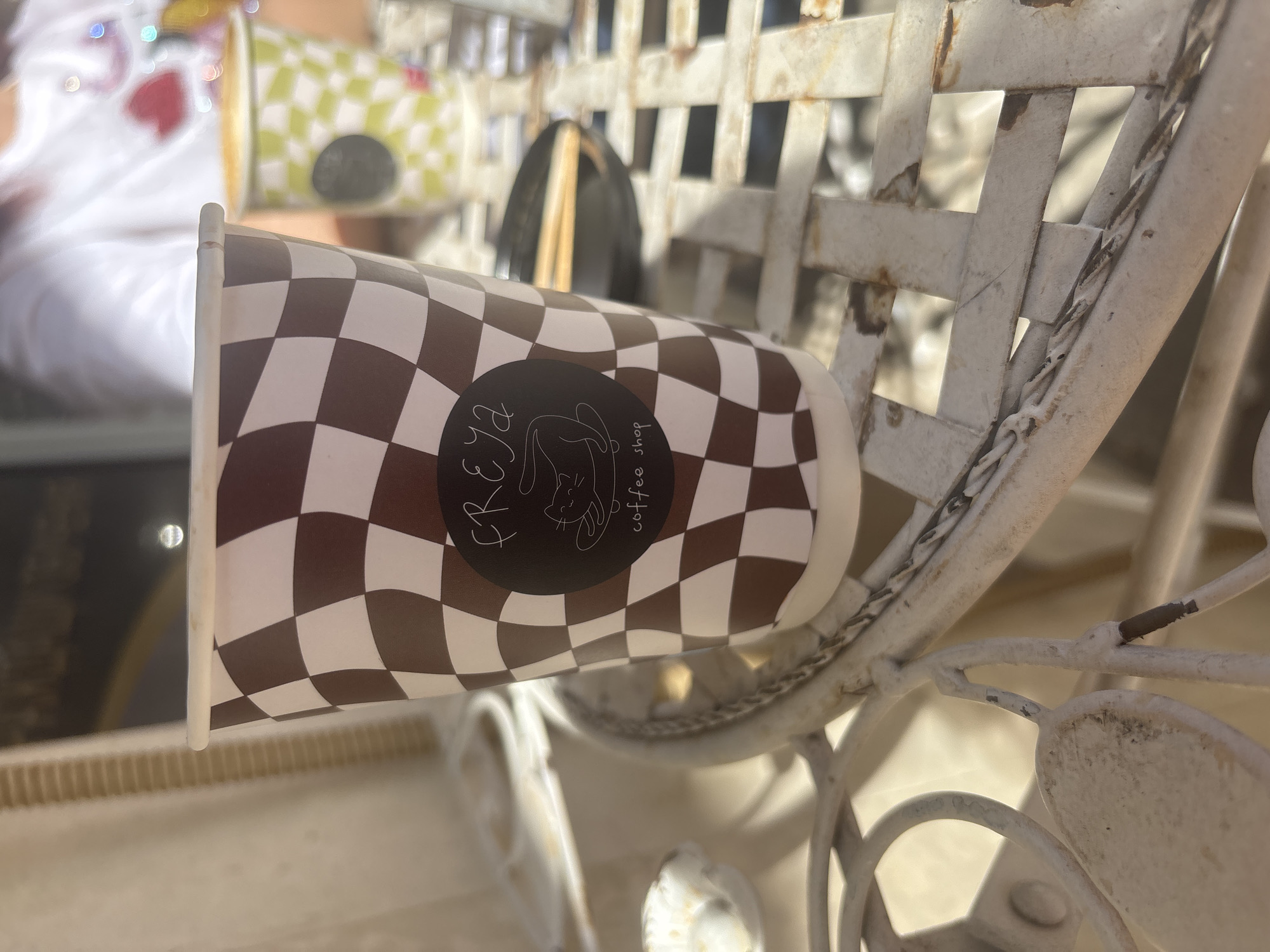 The image captures a close-up of a coffee cup leaning against an ornate, weathered white metal chair, likely an outdoor or patio setting. The paper coffee cup features a distinctive brown and white checkered pattern. A circular black label on the cup prominently displays white text that reads "KREYA coffee shop," accompanied by a stylized white line-art illustration of a cat.
The metal chair shows visible signs of age and wear, with chipped paint and rusty spots, suggesting it's an older piece of furniture. In the blurred background, another person is partially visible, wearing a white garment adorned with colorful, sparkling elements and a red heart shape. Another checkered coffee cup, this one in green and white, is also out of focus in the background near the person.
The scene is well-lit by natural light, indicating it is daytime. The shallow depth of field keeps the foreground cup sharp while blurring the background elements. There is no explicit activity depicted, but the presence of coffee cups suggests a moment of rest or social interaction at a coffee shop. Based on the provided context, the location is Sofia, Bulgaria.