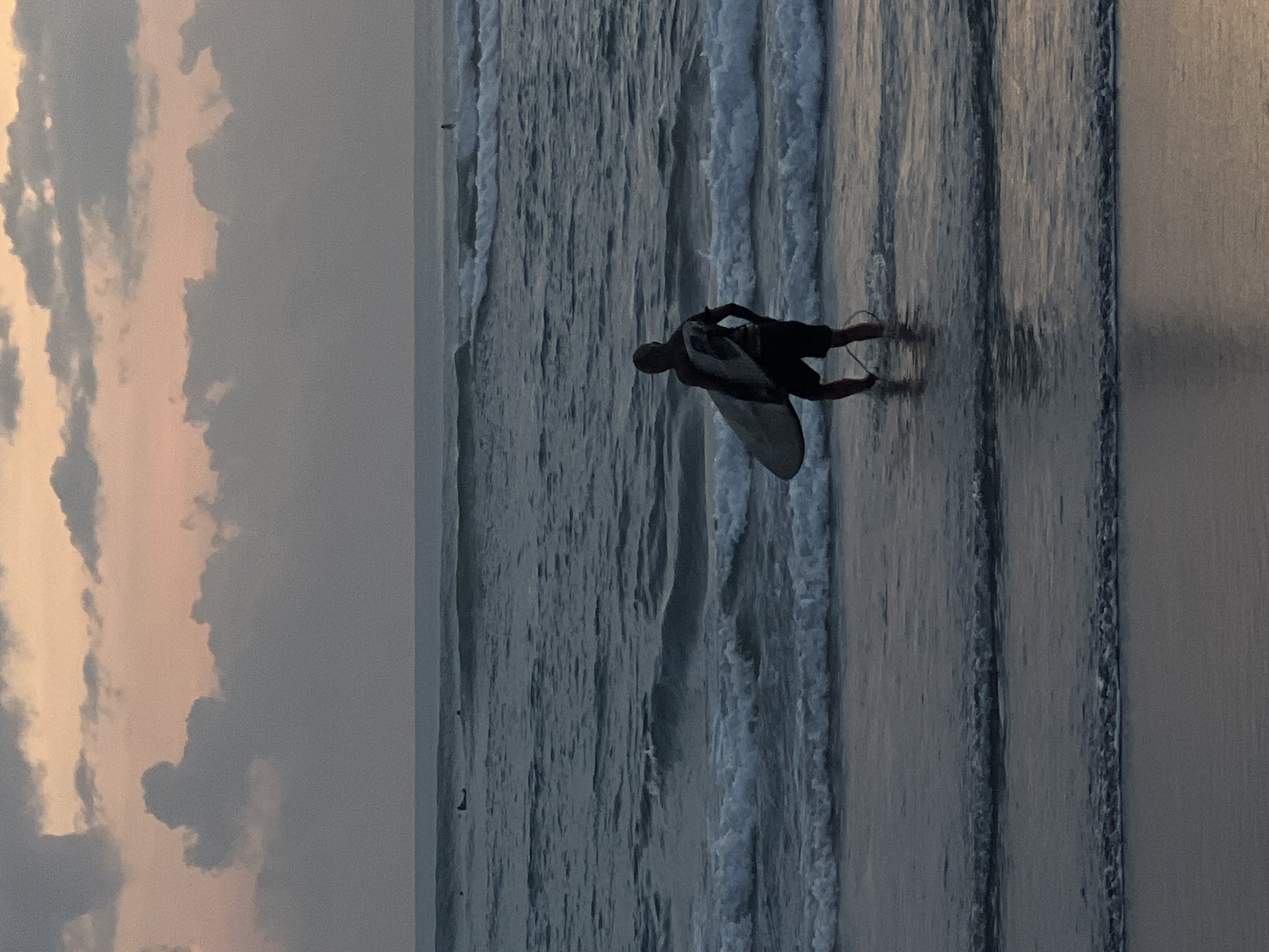 A surfer is seen waist-deep in the ocean at Canggu, Indonesia, carrying a surfboard towards the incoming waves. The scene captures a cloudy sky.