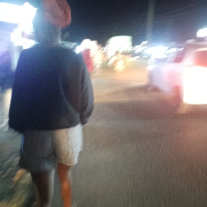 A blurred image depicts a person from behind, wearing a dark jacket and light-colored shorts, possibly walking on a street at night. Light sources from vehicles and buildings are visible in the background, adding to the blur and indistinctness of the scene. The overall impression is that of a snapshot taken in low light conditions with a moving camera.