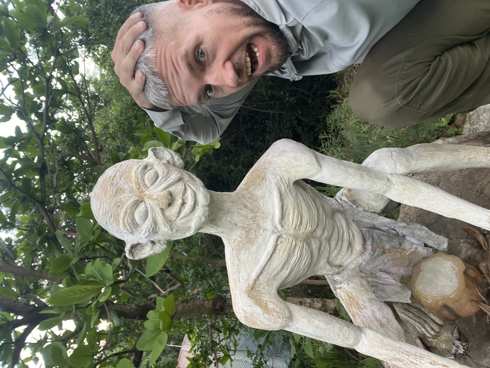 The image displays a male individual posing next to a pale, emaciated humanoid statue resembling "Gollum" from *The Lord of the Rings*, set within a lush, green outdoor environment in Nha Trang, Vietnam.
The male subject, appearing Caucasian, has short, faded grey hair and a short grey beard. He wears a light grey hooded top and dark olive-green pants. He is crouching or kneeling to the right of the statue, facing the camera with an exaggerated wide-eyed, open-mouthed expression of surprise, his left hand gripping the top of his head.
The statue, made of a light-colored material with off-white and subtle brown tones, depicts a skeletal figure with prominent ribs, visible musculature, long limbs, and large ears. Its face features closed or sunken eyes, deep wrinkles, and a wide, toothy smile. It is in a crouching position on a natural or imitation rock base. Between the statue's legs rests a small, spherical object, possibly a painted coconut or rock. The statue exhibits minor surface wear and cracks.
The background consists of dense, green tropical foliage, including trees with broad leaves and various shrubs. A light-colored, possibly corrugated metal, structure is partially visible in the upper left background. The ground is covered with natural vegetation and debris. The scene is illuminated by diffused light, suggesting an overcast day or filtered sunlight.