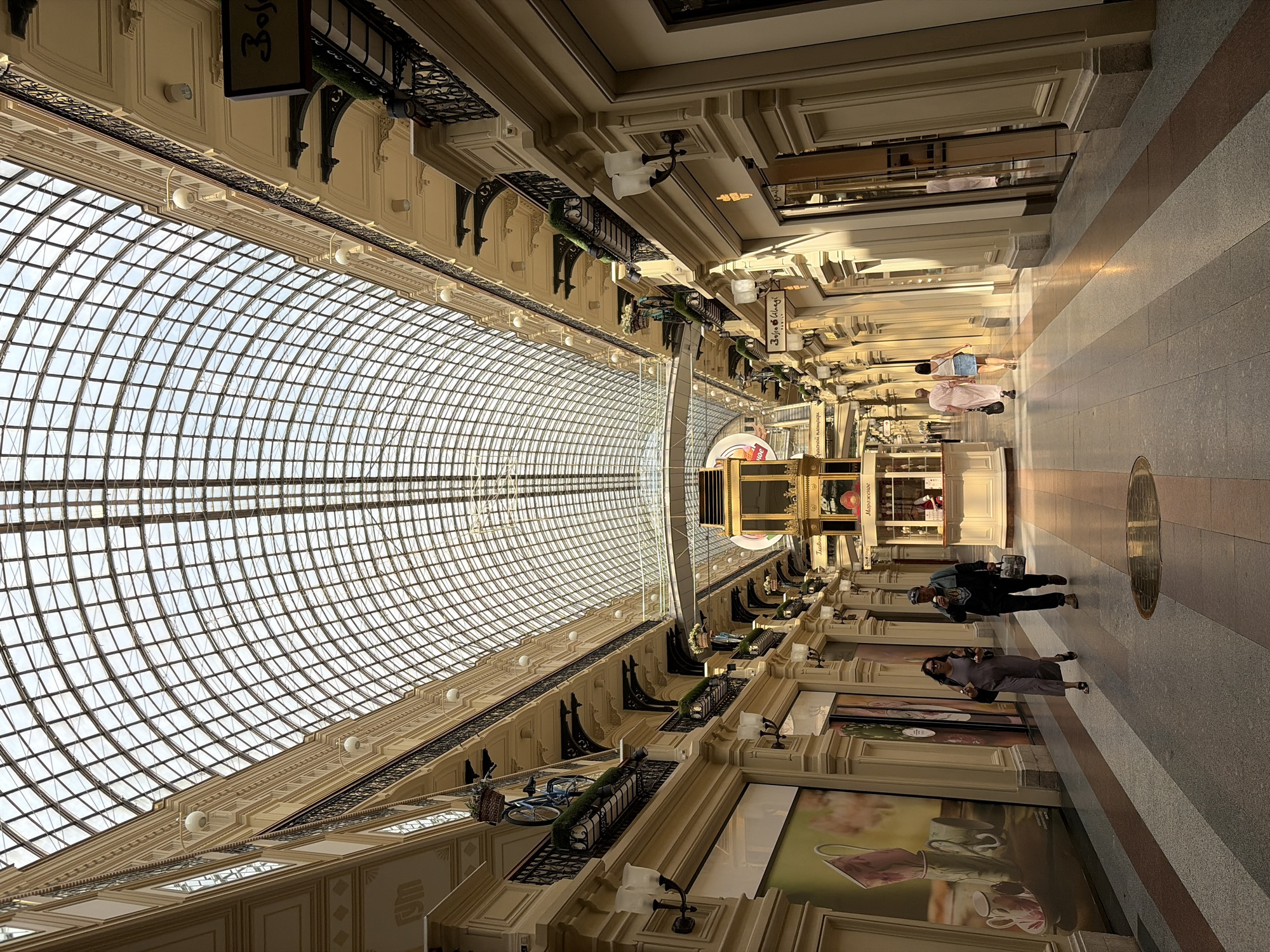 The image depicts the interior of the GUM department store on Red Square in Moscow, Russia. The long, multi-story building features a distinctive glass-vaulted roof that arches high above the main arcade. Natural light streams through the glass ceiling, illuminating the polished floor and the shops lining both sides of the walkway. The architecture is ornate, with classical columns, decorative balconies, and intricate metalwork.
In the foreground, several people are walking along the arcade. On the left, a woman in a gray jumpsuit is walking away from the camera, carrying a bag. To her right, a man in a dark jacket and cap is walking towards the camera, holding a takeaway drink and a bag. Further down the arcade, two women in light-colored dresses are walking away from the camera.
In the middle of the arcade, there is a decorative kiosk with a golden facade, which appears to be a kiosk selling ice cream or some other treat. There are also several storefronts visible, some with large advertisements or displays. The floor is paved with a patterned tile in shades of gray, beige, and brown. The overall impression is of a bustling, elegant, and well-lit shopping environment.