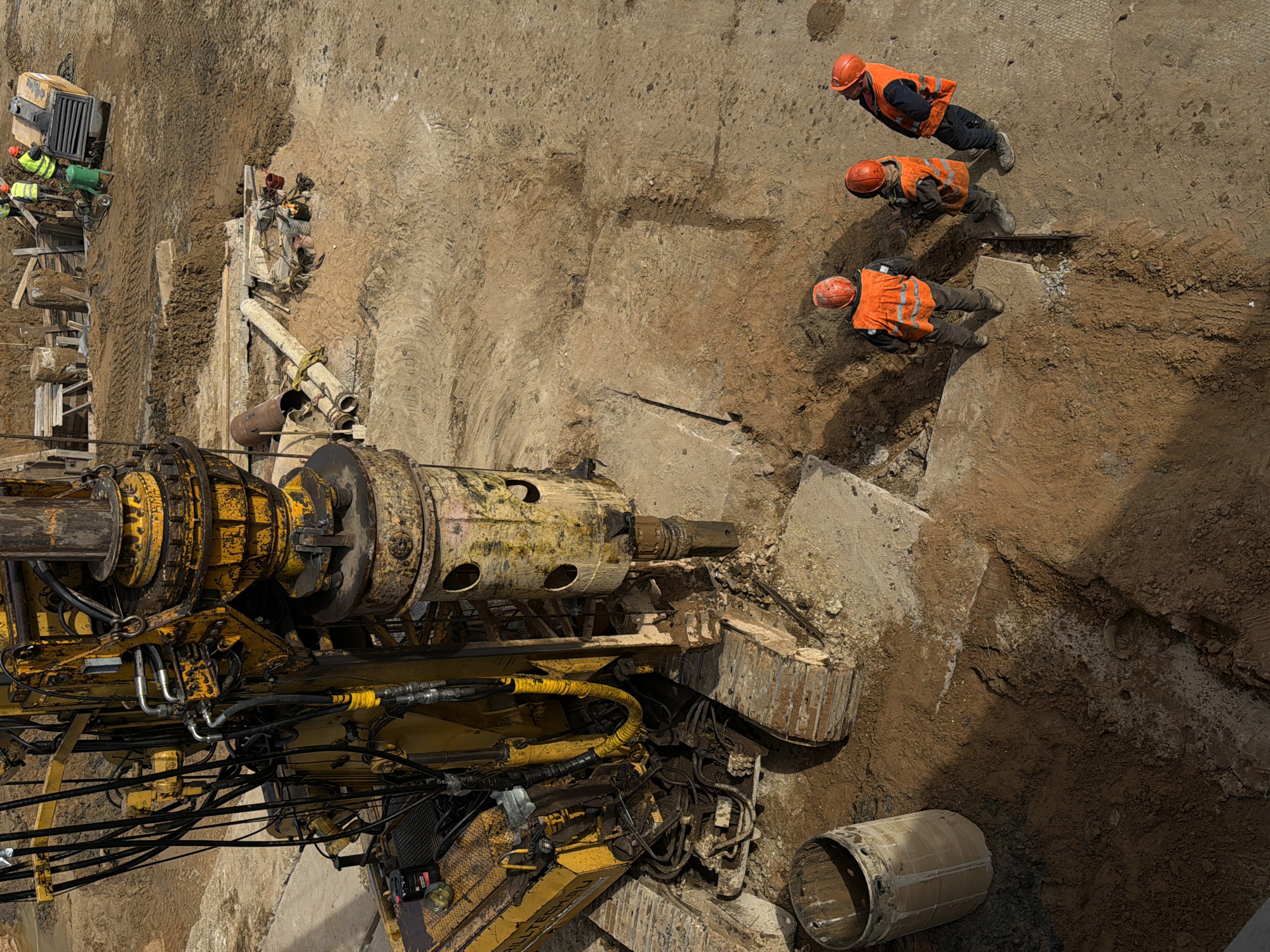 The image shows a construction site, possibly in Moscow, Russia, from an elevated perspective. Three workers wearing orange safety vests and hard hats are positioned on the right side of a deep excavation. A large, yellow drilling machine occupies the lower left portion of the frame. To the left, workers in lime vests are next to a section with wood and other equipment. A cylindrical metal section is next to the drilling machine. The surrounding environment is primarily composed of earth and concrete. The construction area seems to be a major excavation project.