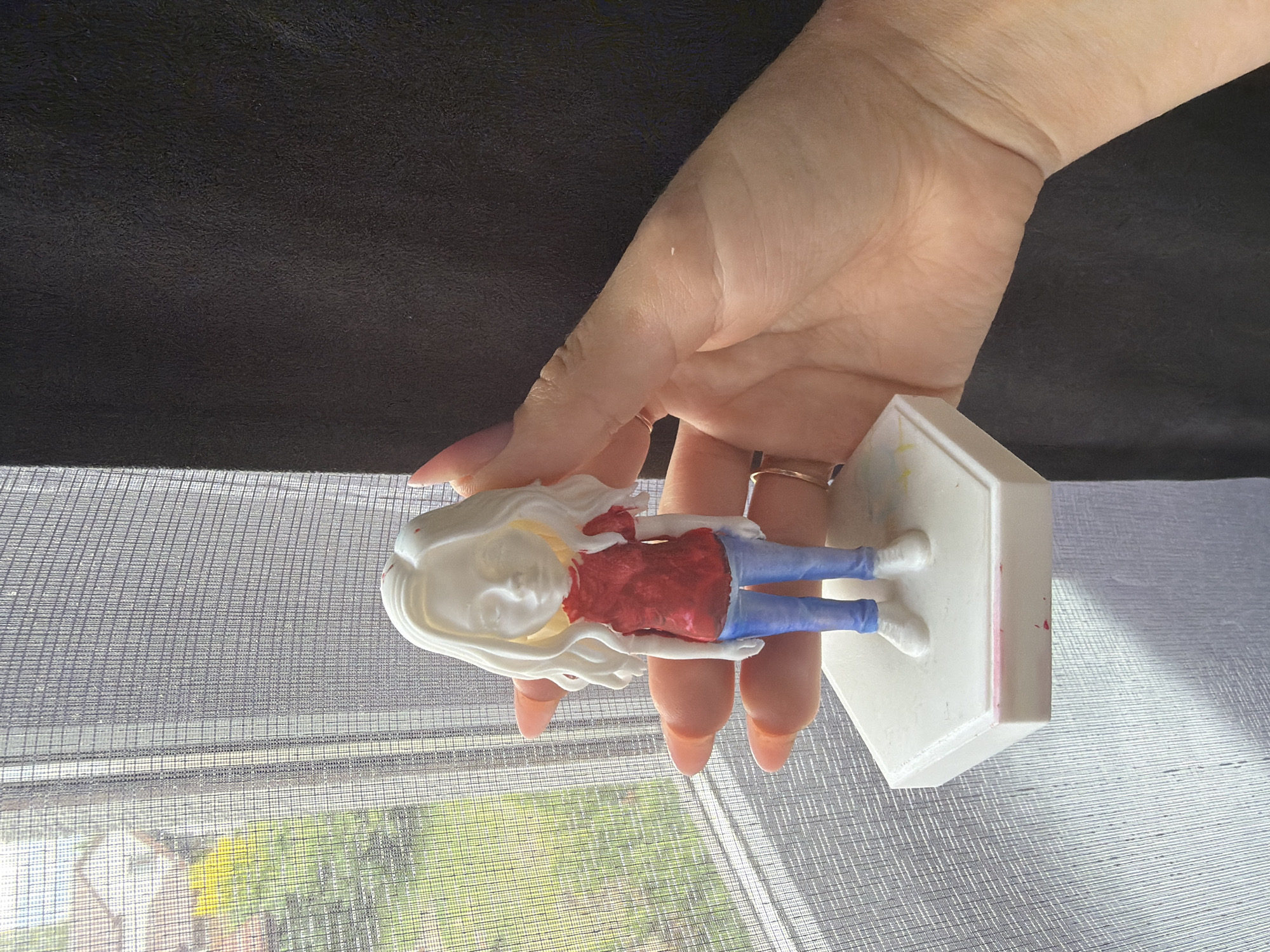 A right hand, with light peach-colored manicured fingernails and a gold-toned ring on the ring finger, holds a white, unpainted figurine. The figurine depicts a female figure with long, wavy, unpainted white hair, an unpainted blank face, a textured red short-sleeved top, light blue jeans, and white shoes. It stands on a white hexagonal base, which has minor red and yellow paint smudges. The background on the left consists of a window covered by a fine mesh screen, revealing outdoor scenery with vibrant green foliage, yellow flowers, and a distant building under natural light. To the right of the hand, a dark, textured surface, possibly fabric, is visible. The scene is illuminated by light entering through the window.