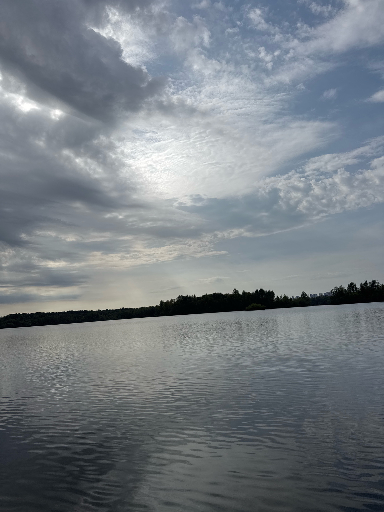 This is a photograph taken from a body of water, likely a lake or river, looking towards a shoreline with trees and a distant cityscape. The sky is filled with dramatic clouds, with patches of blue sky visible. The water is calm with ripples reflecting the sky. The environment appears to be natural with a touch of urban development on the horizon. The image does not contain any people or animals. Based on the visual cues and the provided information, the location is likely the city of Ufa, Russia. The scene is serene and captures a moment of natural beauty with a hint of civilization.