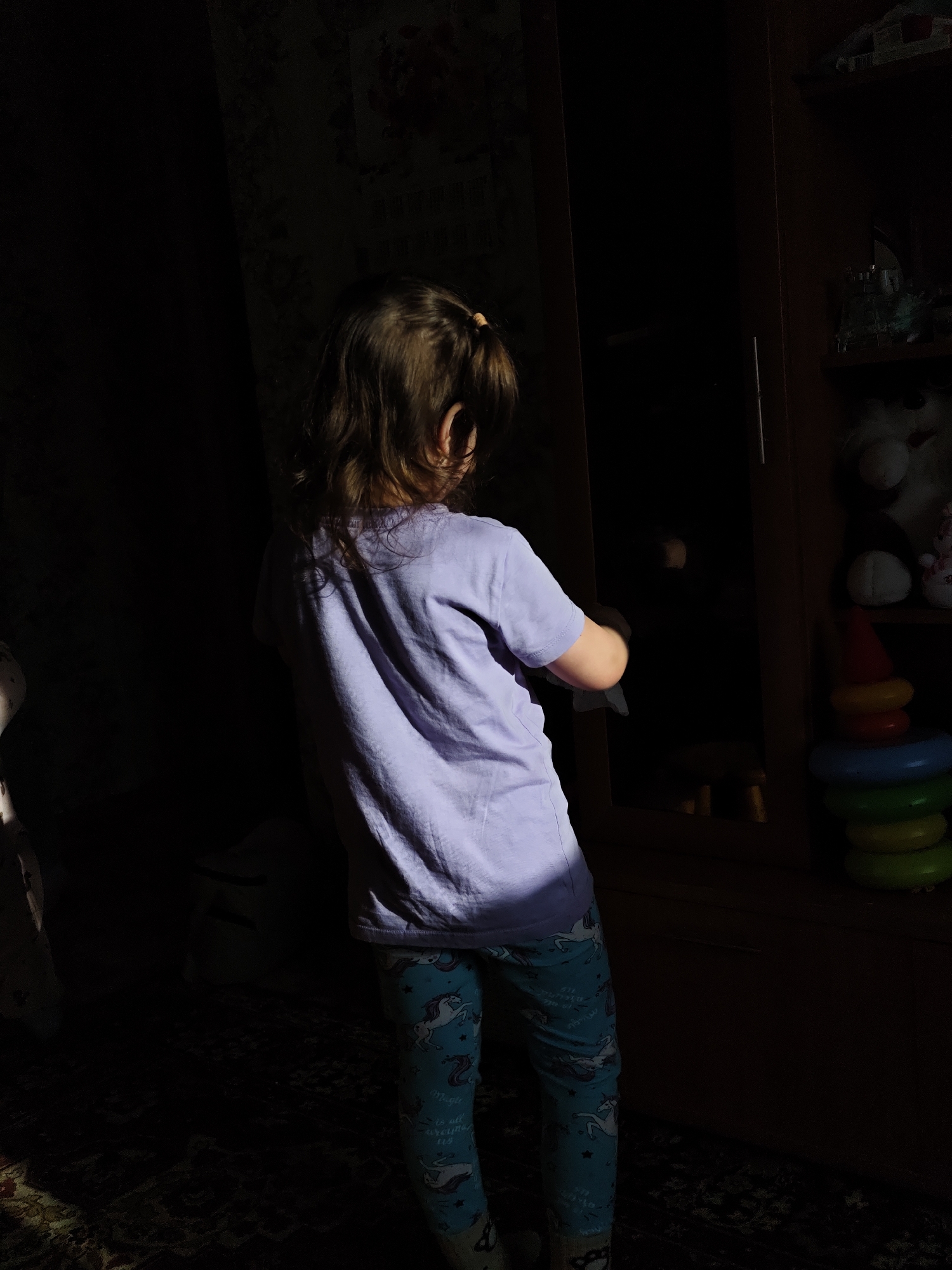 A child stands in a room with a wardrobe in Уральское, Russia. The child, seen from the back, has brown hair tied in a ponytail and wears a light purple t-shirt and blue leggings with unicorn patterns. The child is facing a dark wooden wardrobe that is partially visible. Toys, including stuffed animals and a stacking ring toy, are visible on the shelves of the wardrobe. The room is dimly lit, with a beam of light illuminating the child's torso and legs. A patterned carpet is visible on the floor.