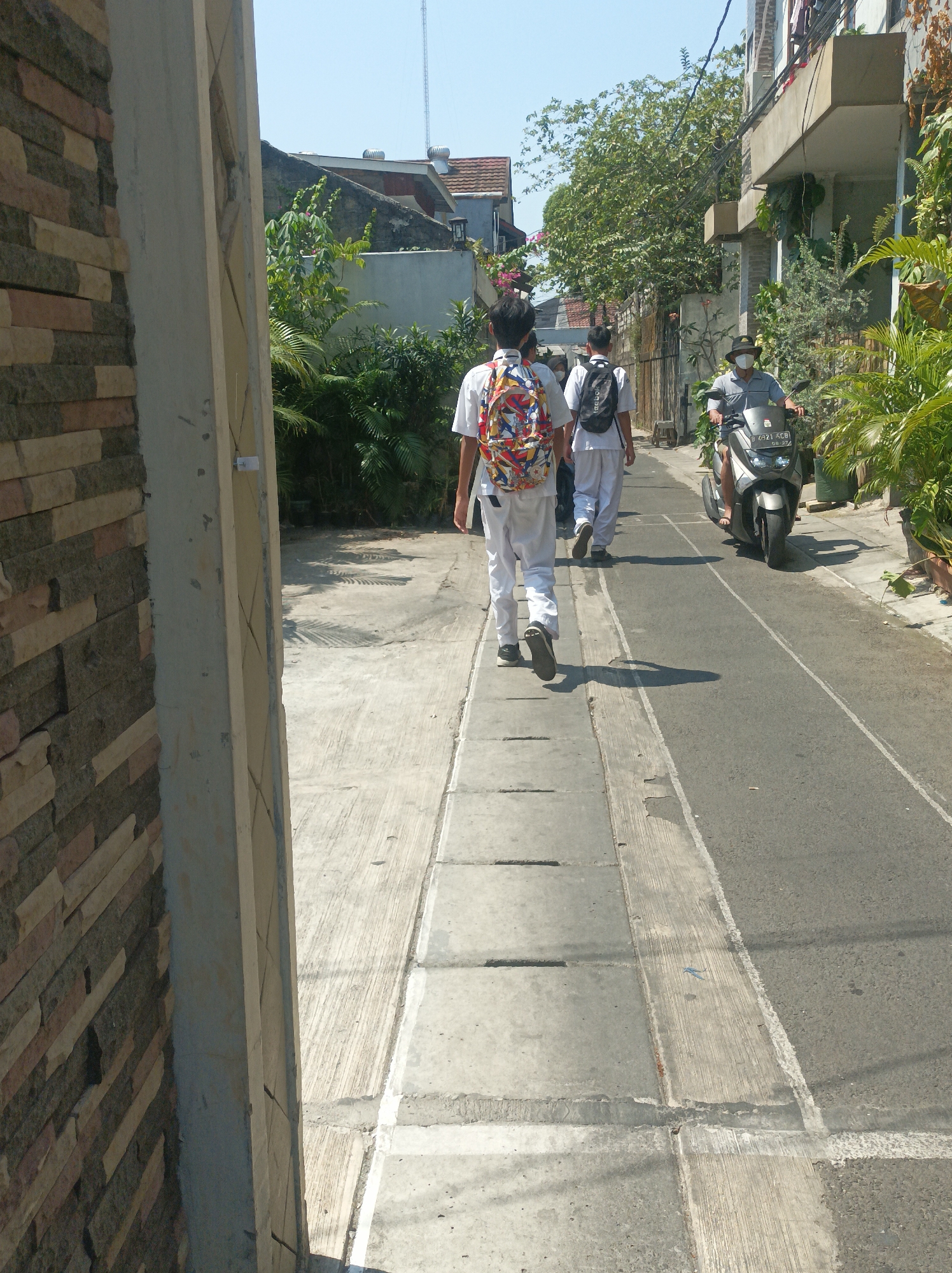 The image depicts a street scene in West Jakarta, Indonesia, on a sunny day. Two students in white uniforms with backpacks are walking away from the camera on a sidewalk. One student has a colorful backpack, while the other has a black backpack. A man wearing a hat and mask is riding a motorcycle on the road alongside the sidewalk. The street is lined with buildings and lush greenery. The sidewalk is made of concrete slabs, and the road is asphalt. The overall impression is one of everyday life in a bustling Indonesian city.