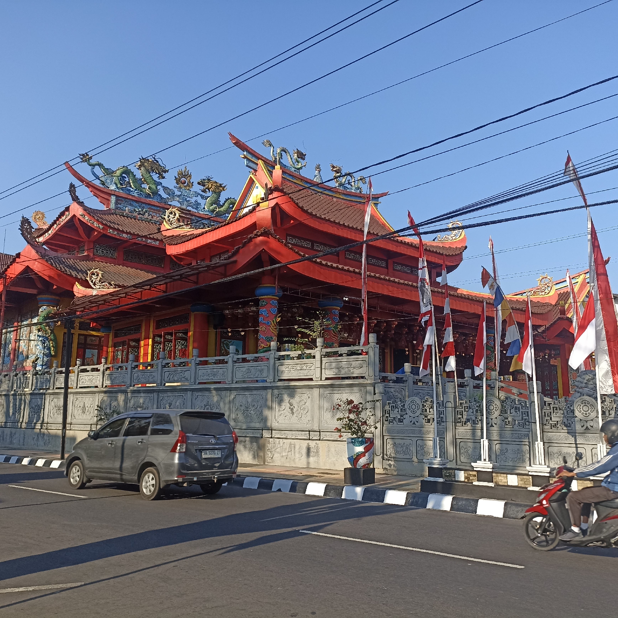 The image shows a temple with red roofs and dragon statues on the roof in Magelang, Indonesia. The temple is decorated with flags and has a grey wall with carvings. There is a grey car parked on the side of the road, and a person riding a red motorcycle on the road. The sky is clear and blue, and the sun is shining. The temple appears to be a place of worship and cultural significance in the city.