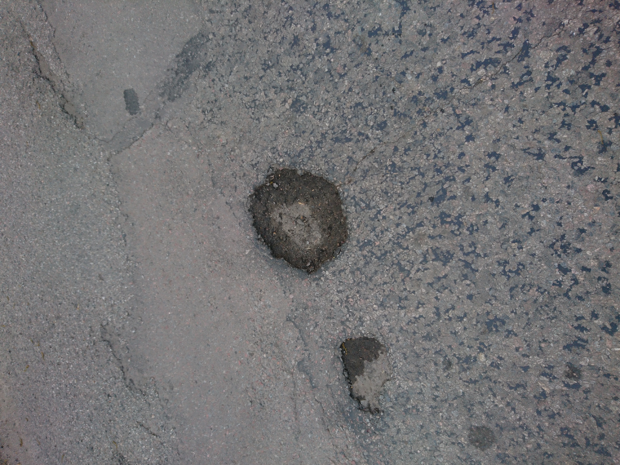 The image shows asphalt pavement in disrepair. There are two distinct patches of darker asphalt that appear to be repairs within the original, weathered asphalt. Cracks and areas of discoloration are visible throughout the pavement. The location is unknown.