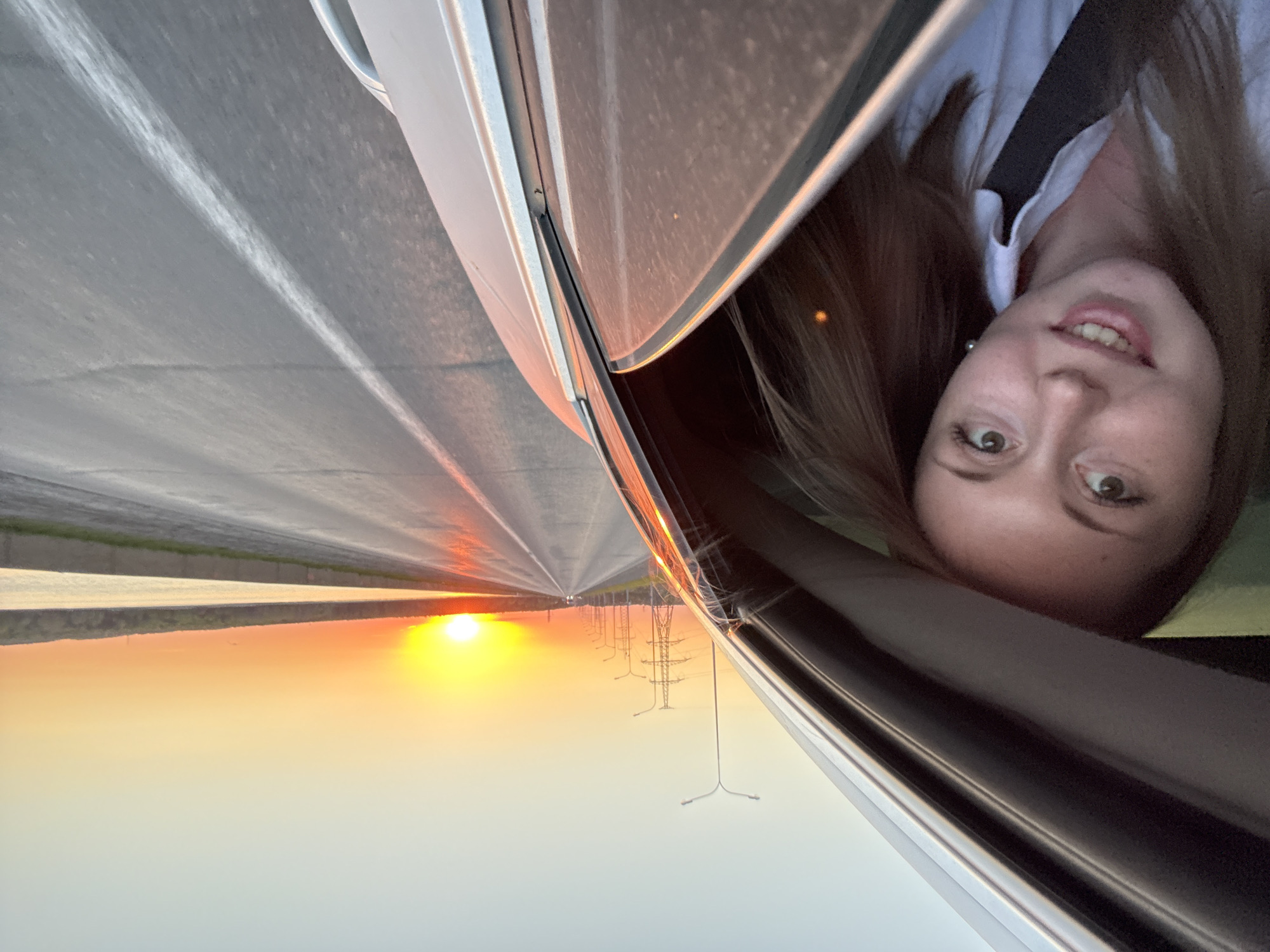 A straight highway extends to the horizon, where a bright sun is visible. Power lines run alongside the road. A woman is looking out of a car window. The location is unknown.