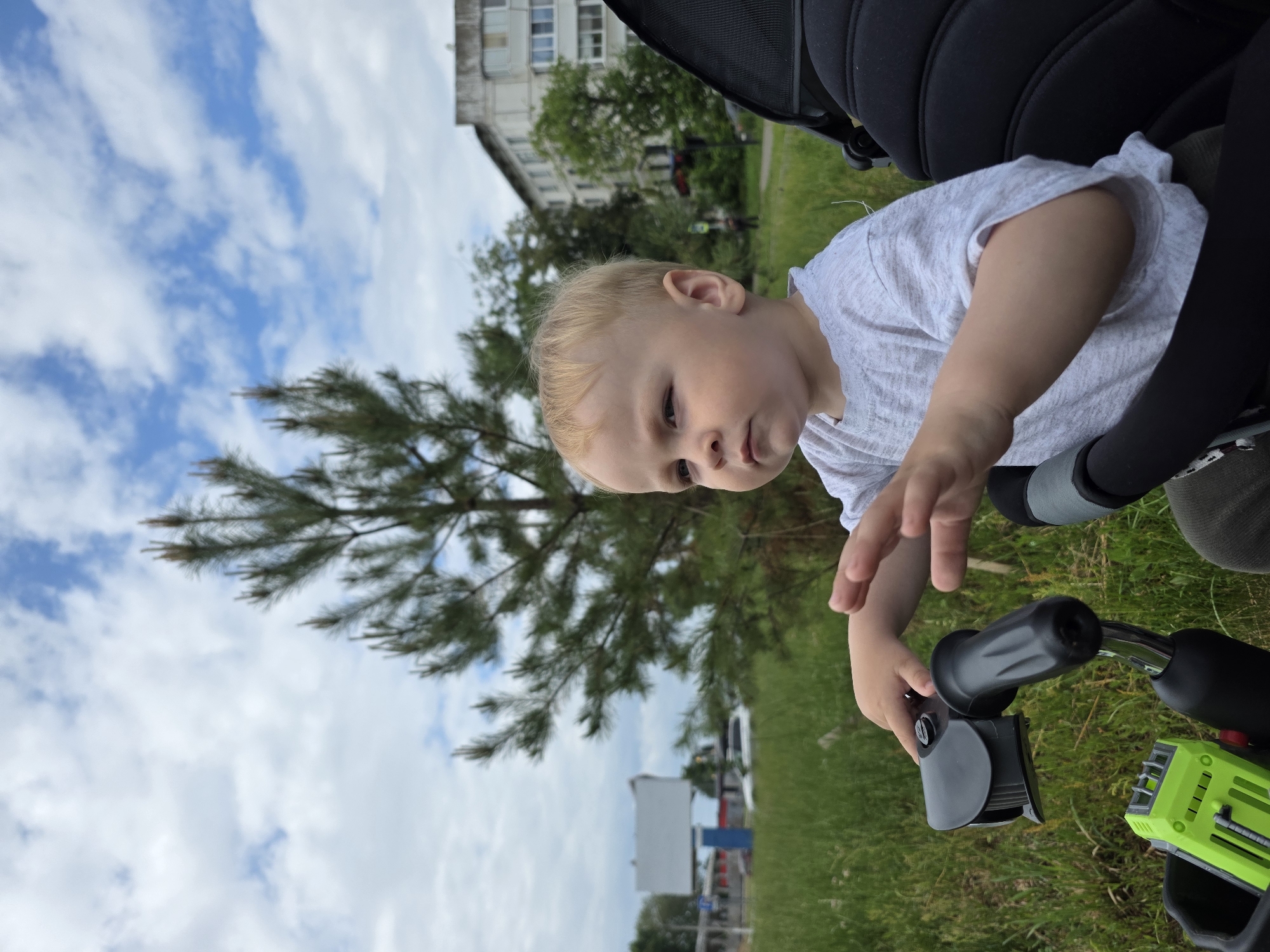 A blonde, light-skinned toddler with blue eyes sits in a black stroller. The child is wearing a grey t-shirt and dark pants. The toddler is looking to the left with a serious expression, with their right hand on the stroller's handle and their left hand in the air. In the foreground, a green toy is visible attached to the stroller. The background shows a large pine tree, a grassy field, a tall apartment building, and a cloudy blue sky. The scene is likely located in a residential area of Chaikovsky, Russia, given the architectural style of the building and the presence of a pine tree, common in the region.